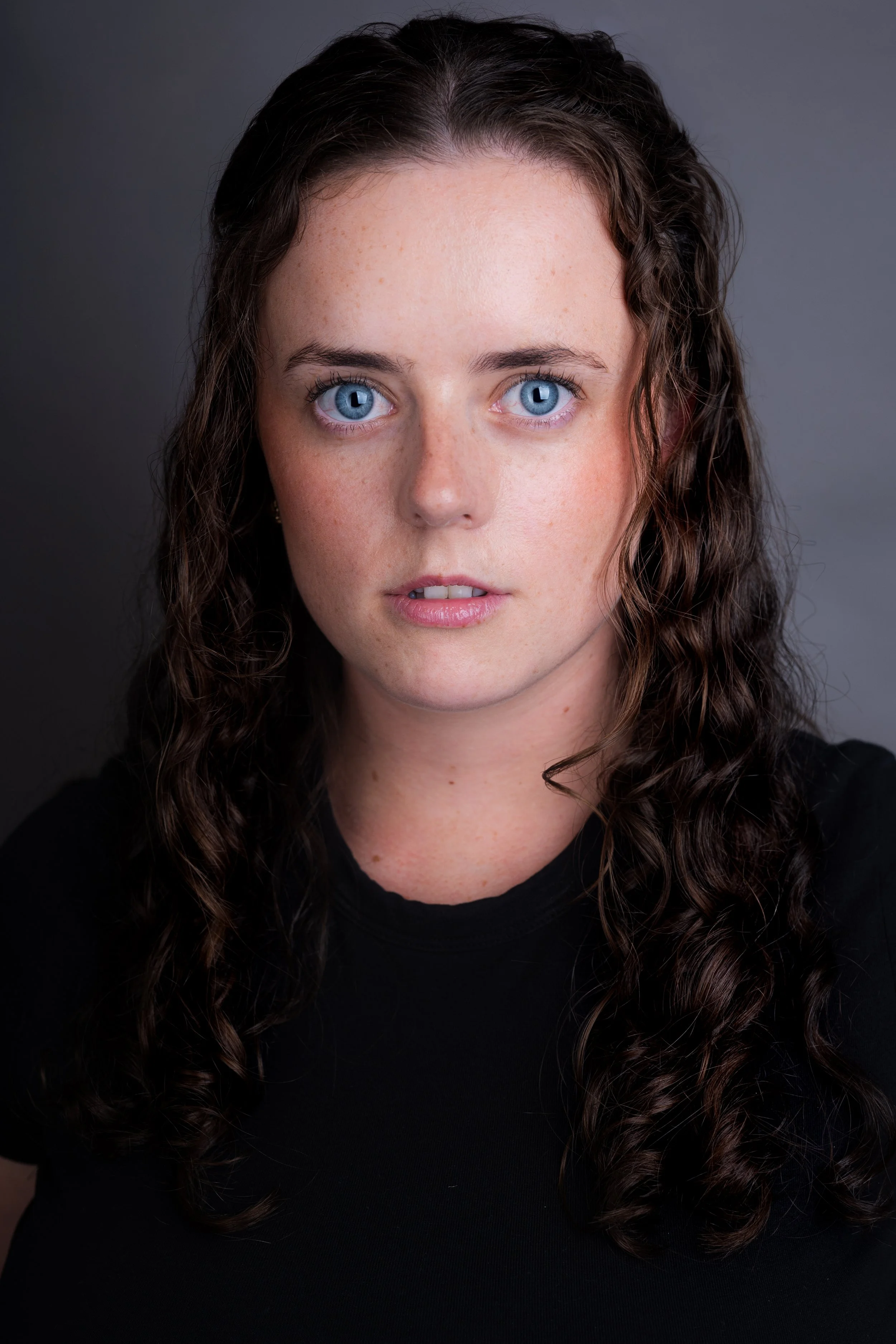Close-up of a woman with long, curly brown hair and striking blue eyes, wearing a black top against a dark background.