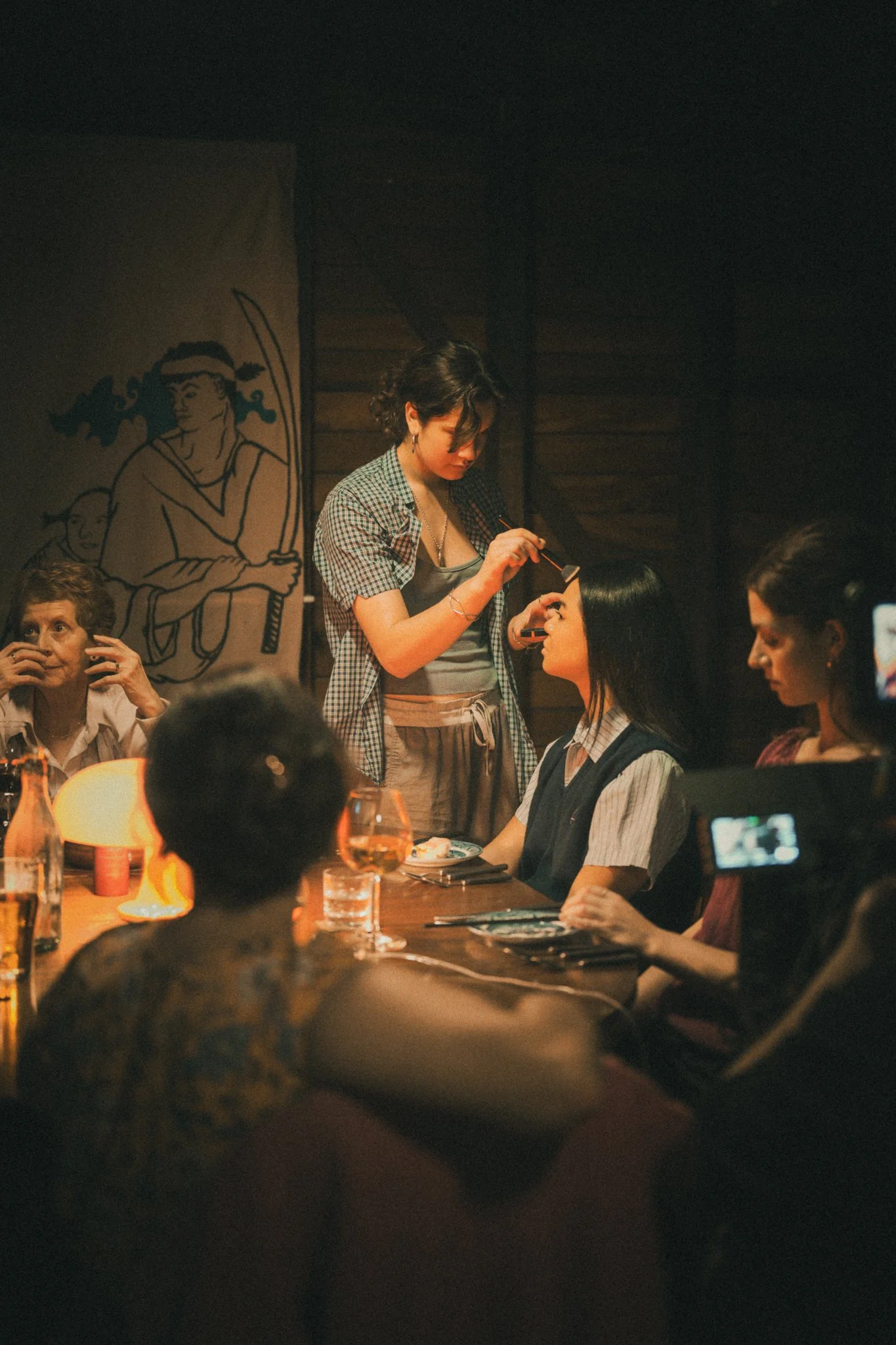 A woman applies makeup to another woman sitting at a table in a dimly lit restaurant or bar with other patrons. There is a large drawing of a woman with long hair on the wall in the background.