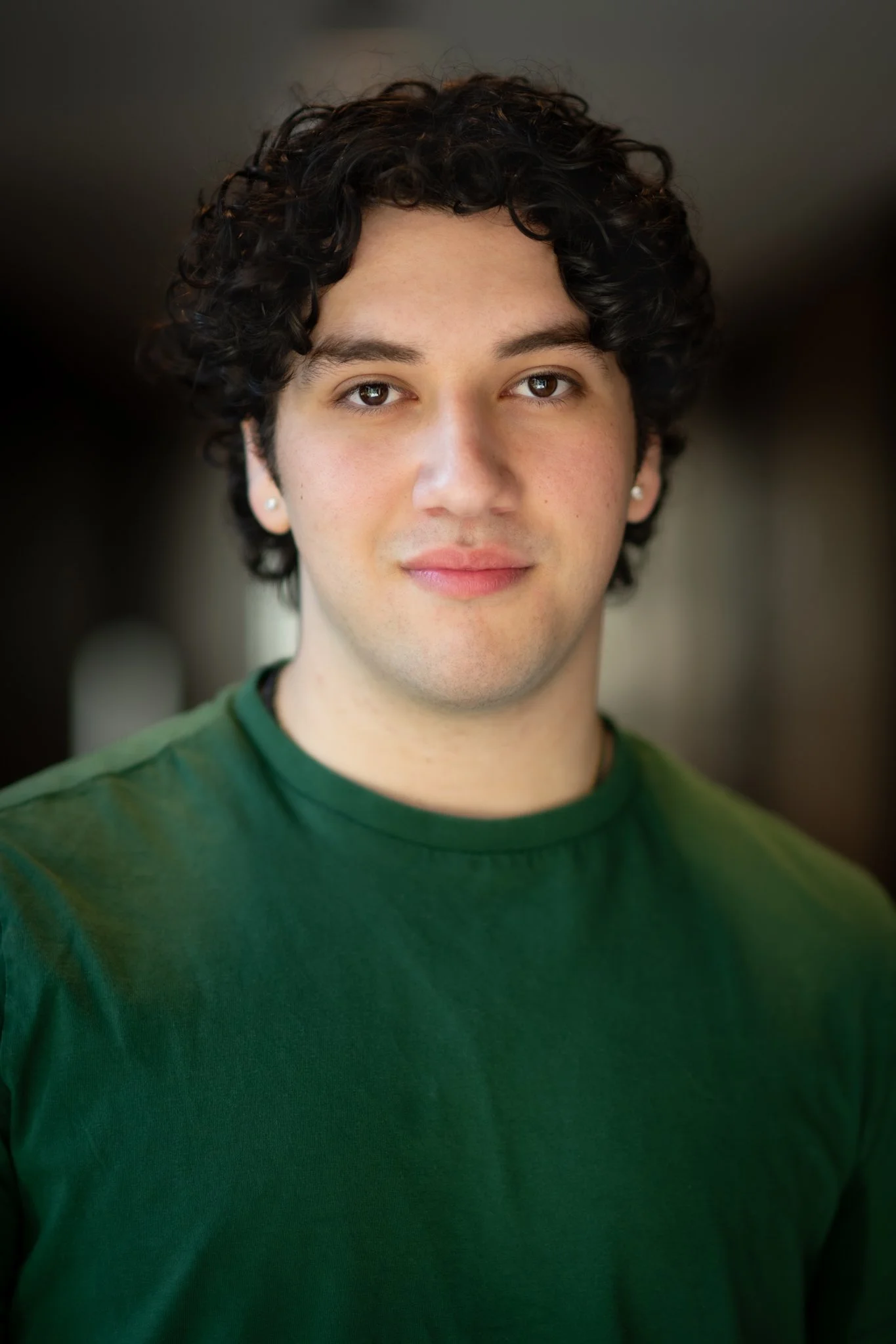 Portrait of a young man with curly dark hair wearing a green shirt and pearl earrings, smiling softly, against a blurred indoor background.