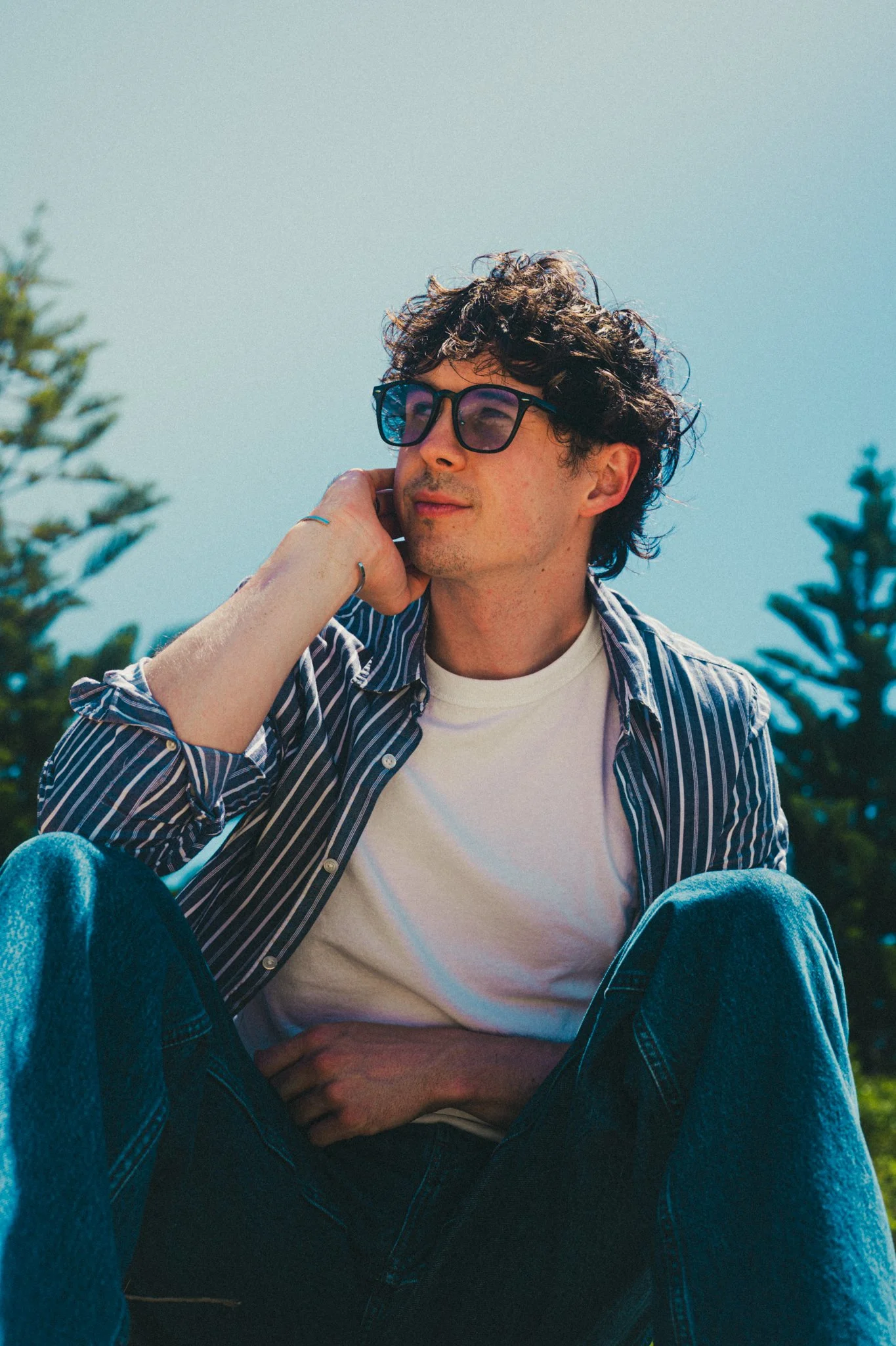 A young man with curly hair and glasses sitting outdoors against a blue sky with trees in the background, resting his head on his hand.