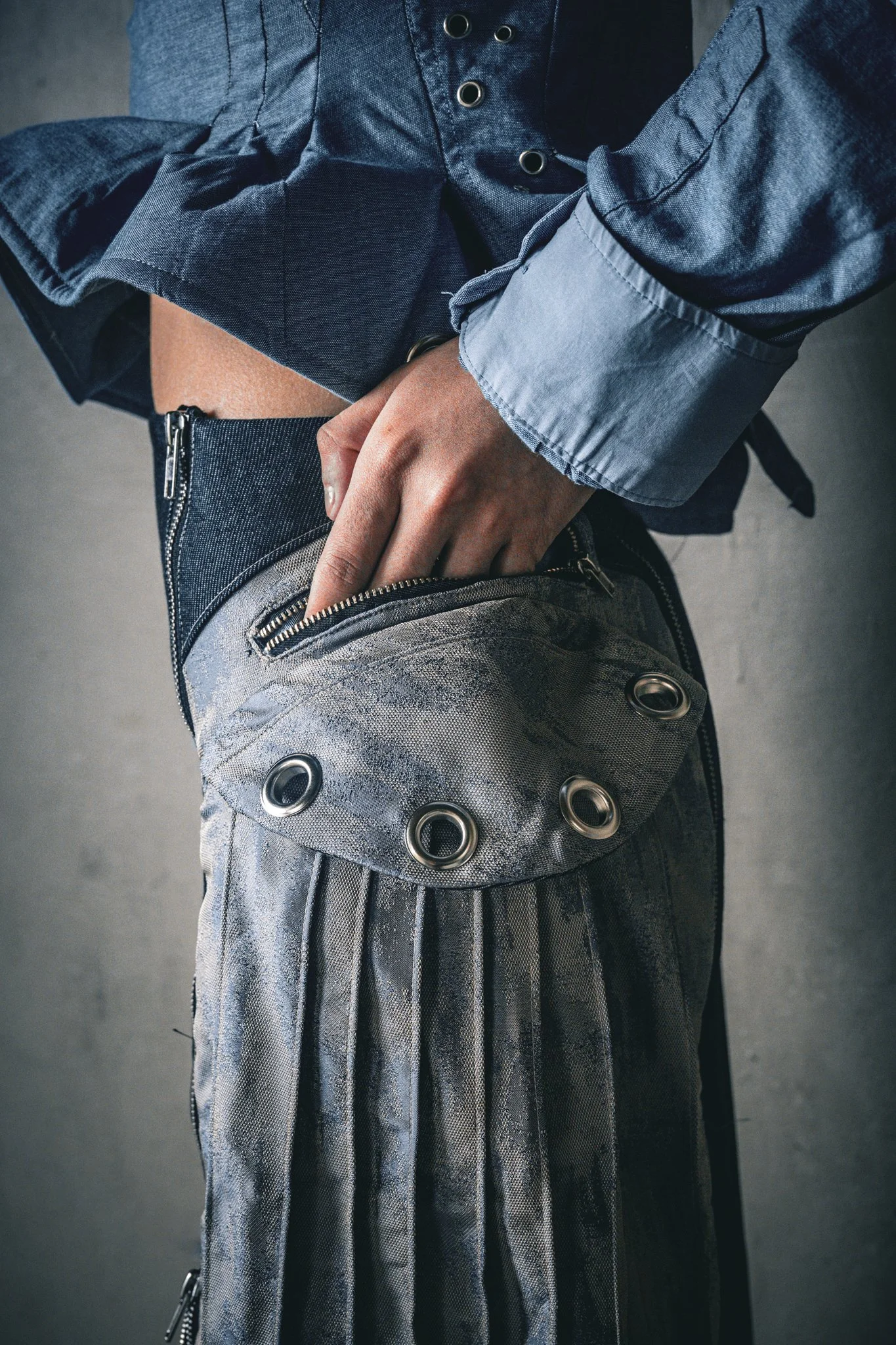 A person wearing a blue shirt and grey plaid skirt with metal eyelet details in a fashion-forward pose.