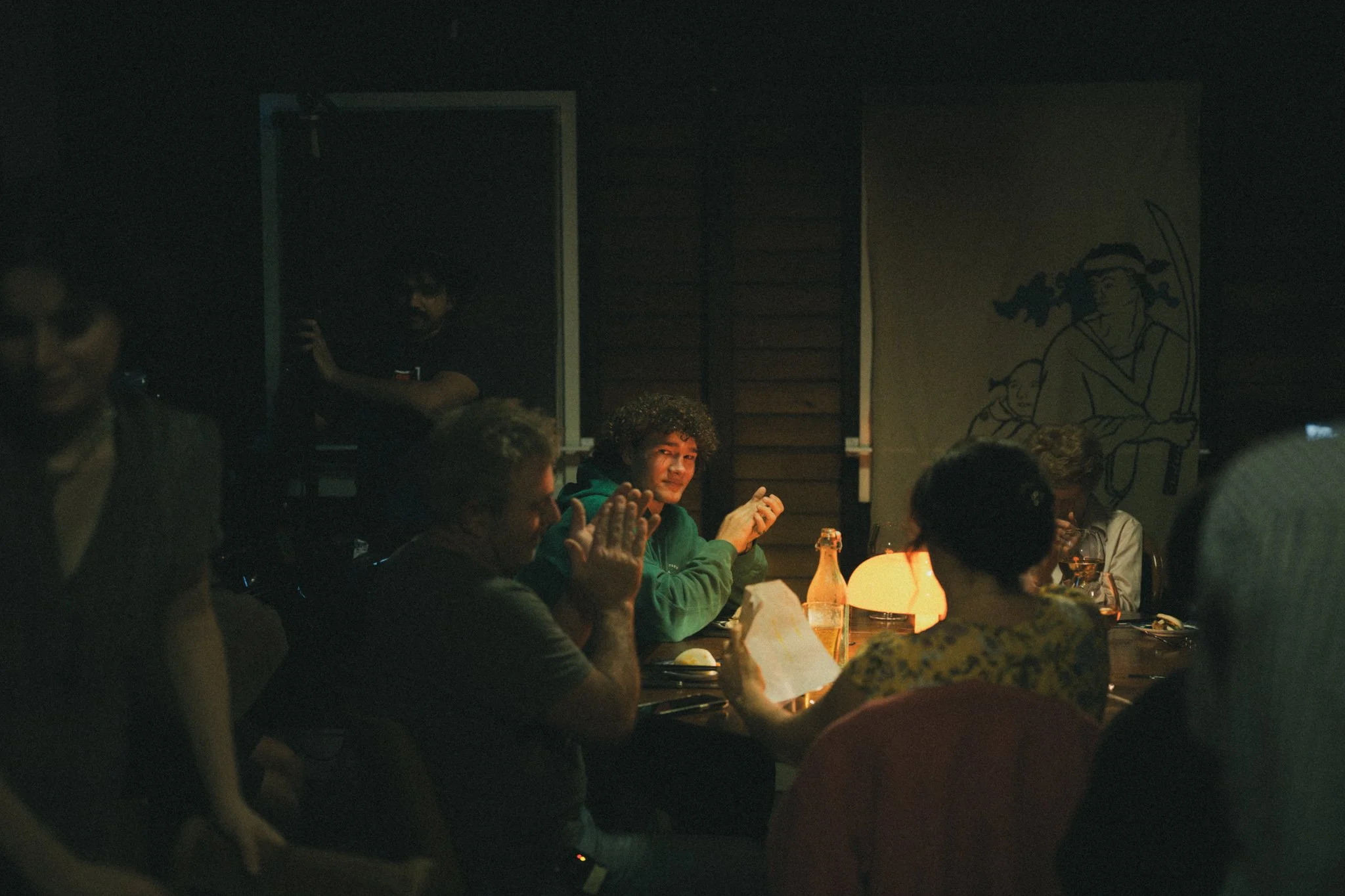 Group of people sitting at a darkened dinner table, with a warm light source illuminating their faces, in a cozy, dimly-lit setting.