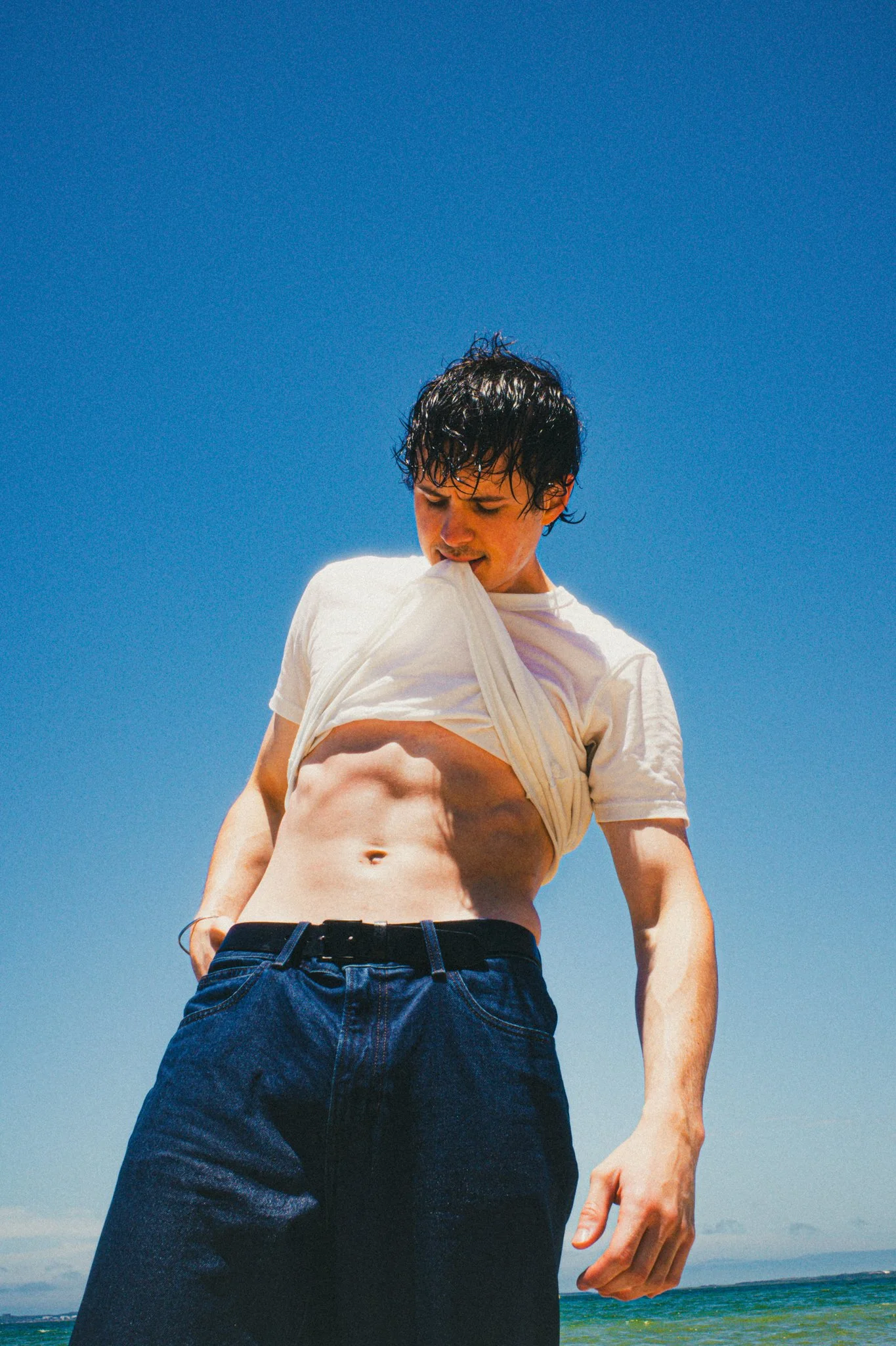 Young man at the beach lifting his shirt to reveal his abs, with clear blue sky and ocean in the background.