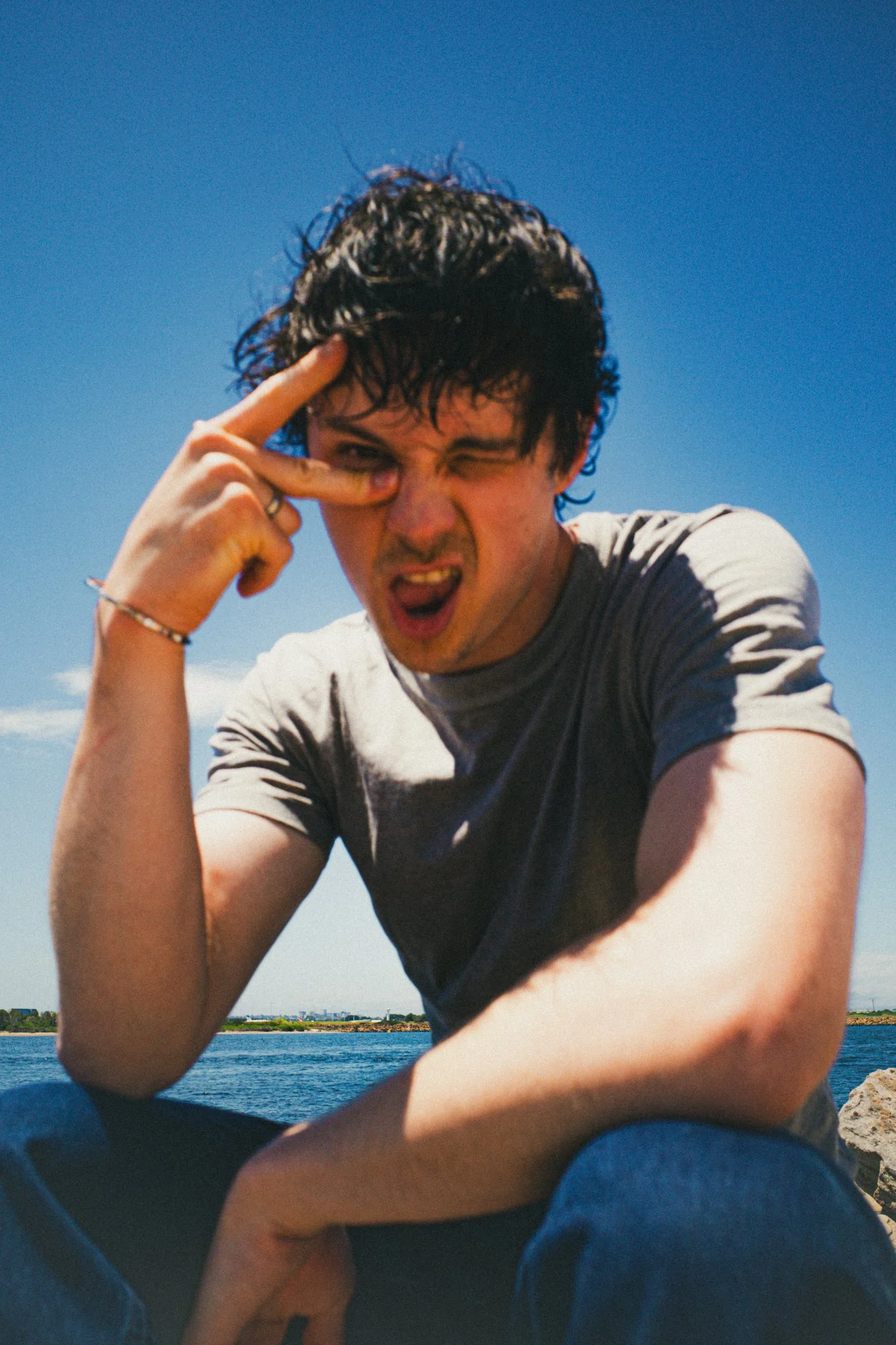 Young man with curly dark hair making a peace sign near his eye, squinting and sticking out his tongue, sitting outdoors near water with clear blue sky.