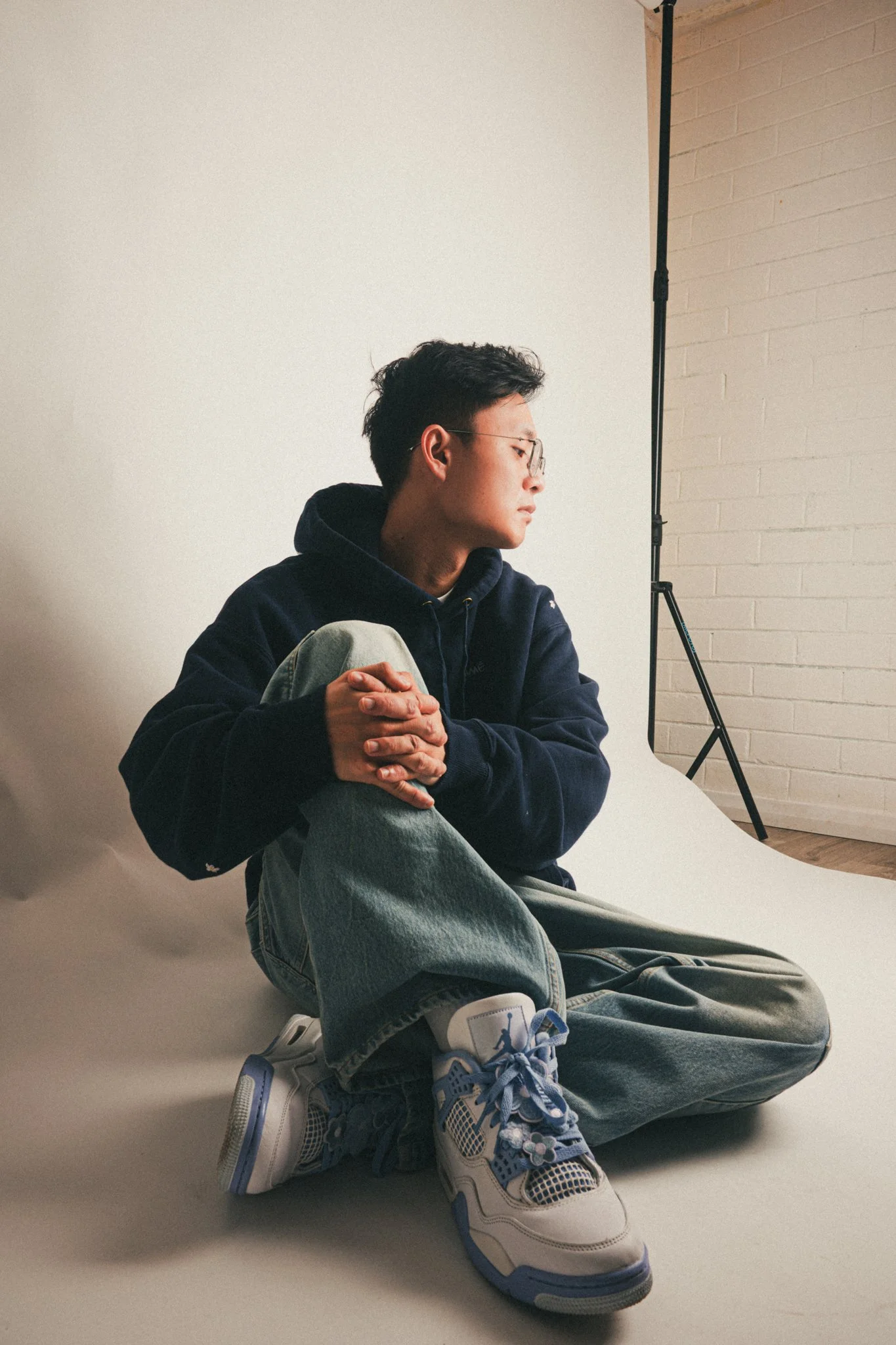Young man sitting on the floor with legs crossed, wearing glasses, a dark hoodie, and sneakers, in a photography studio with a white backdrop and a brick wall.