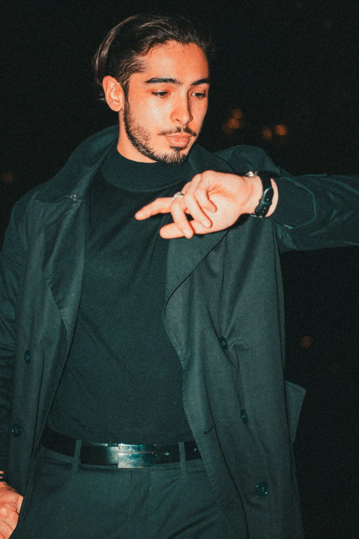 A young man with dark hair and beard wearing a black turtleneck, black coat, and black belt checking the time on his wristwatch amid a dark night background.