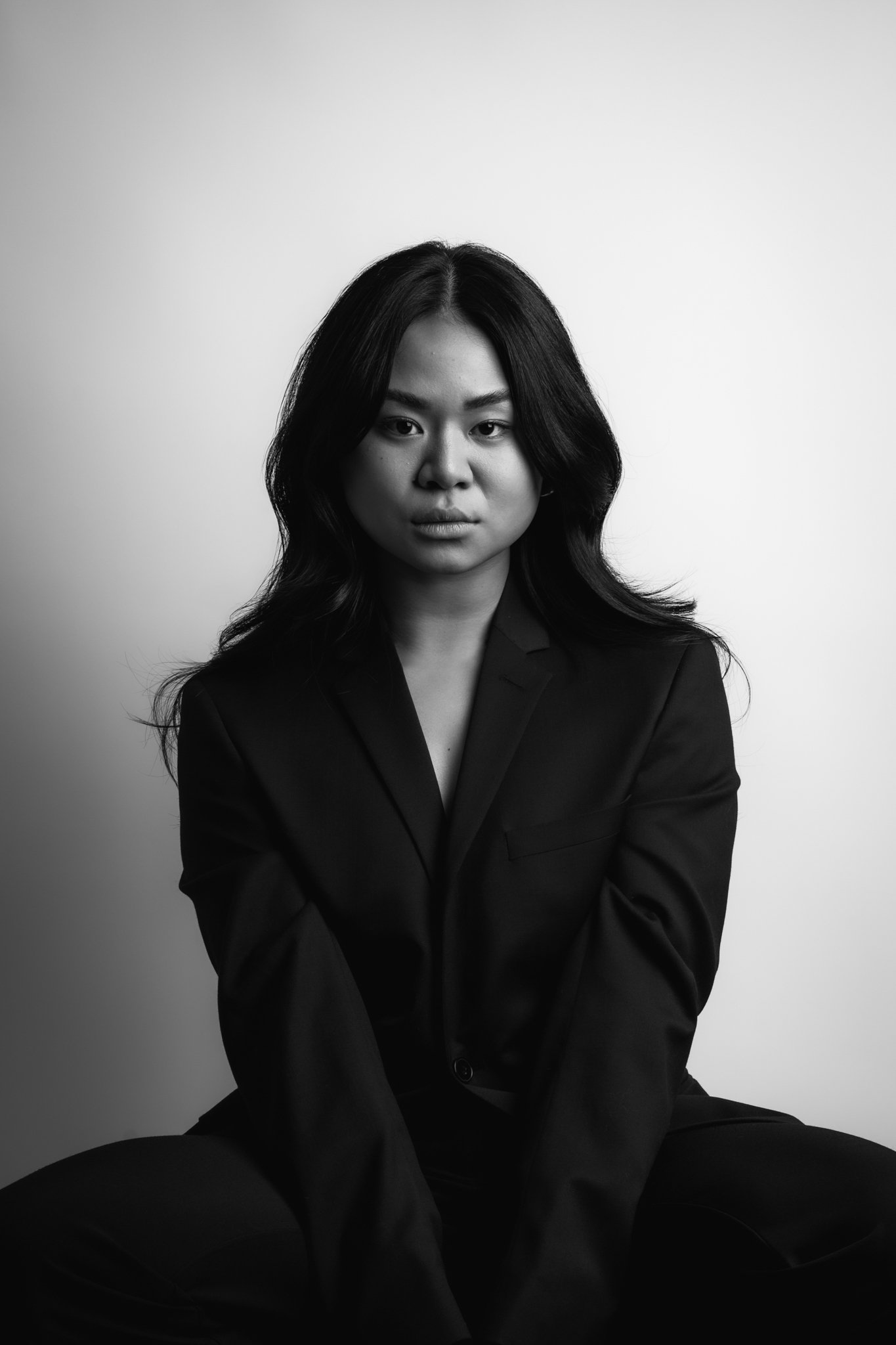 Black and white portrait of a woman with long dark hair wearing a suit, sitting against a light background.
