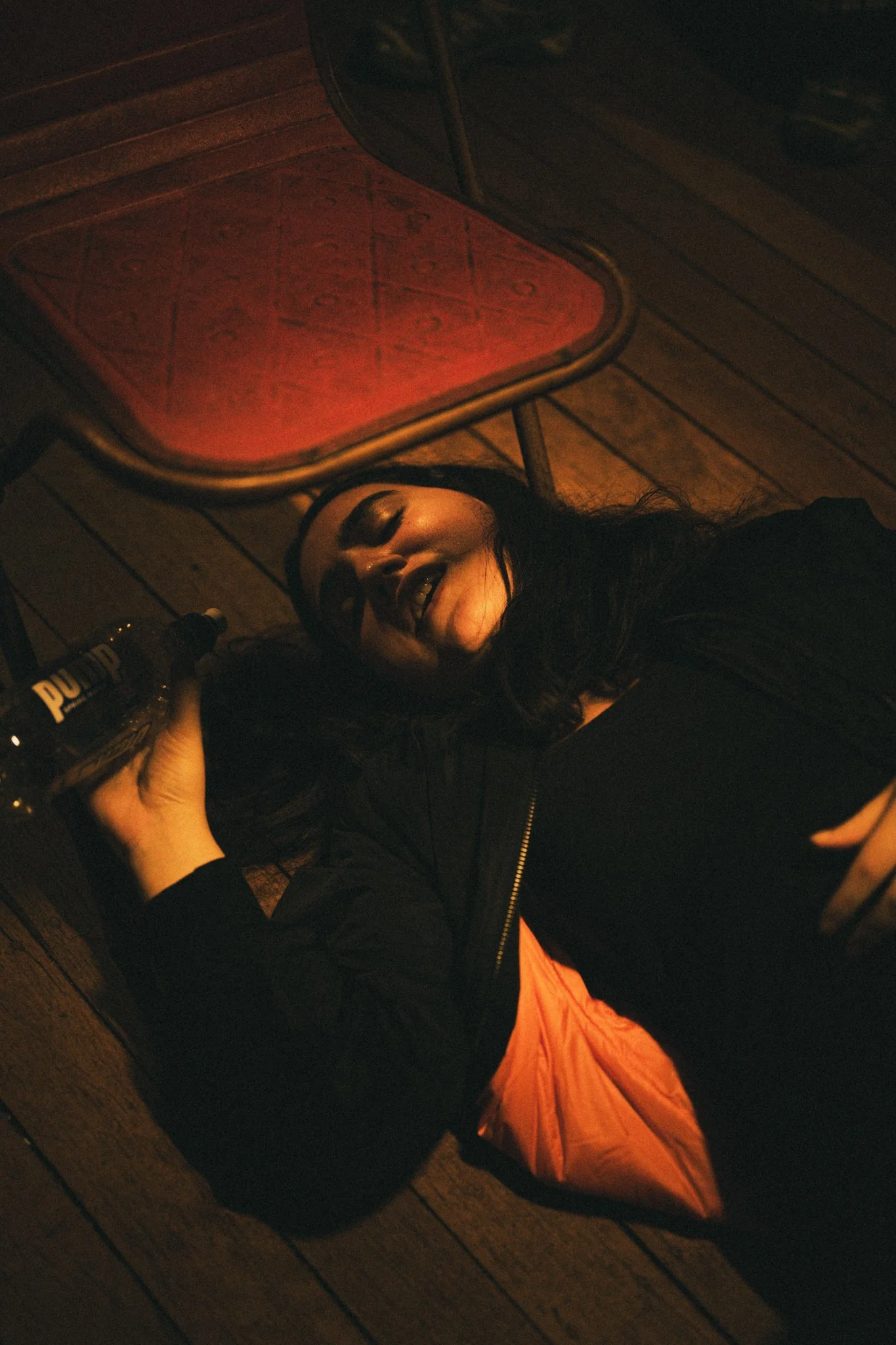 A woman with dark hair lying on the wooden floor, smiling, holding a bottle of Pum and with an orange sleeping bag near her. There is an orange cushion on a small table beside her.