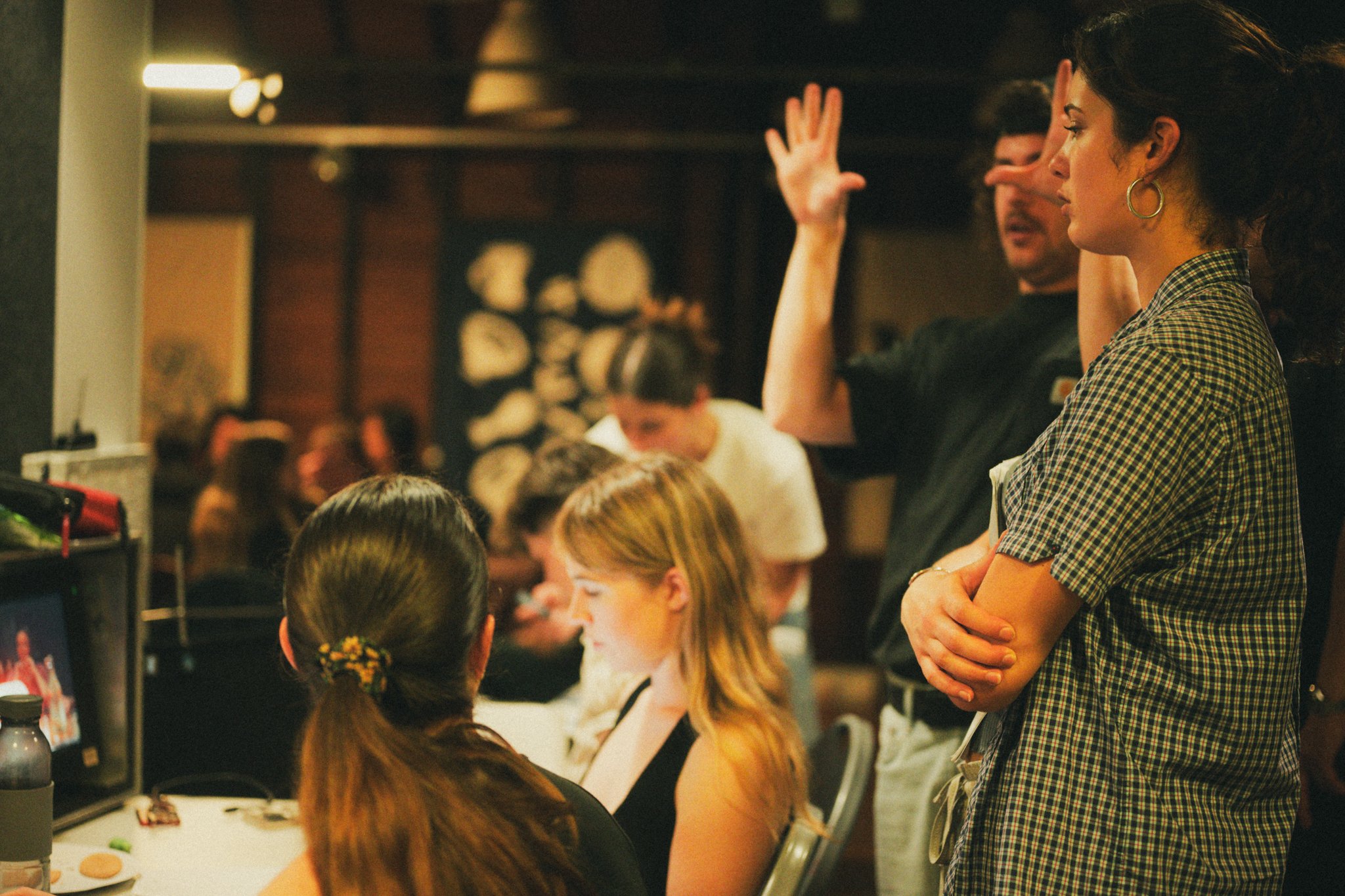 People working on a film or TV set, with a woman in the foreground—standing with her arms crossed and focused—while others are seated at a computer and preparing equipment.