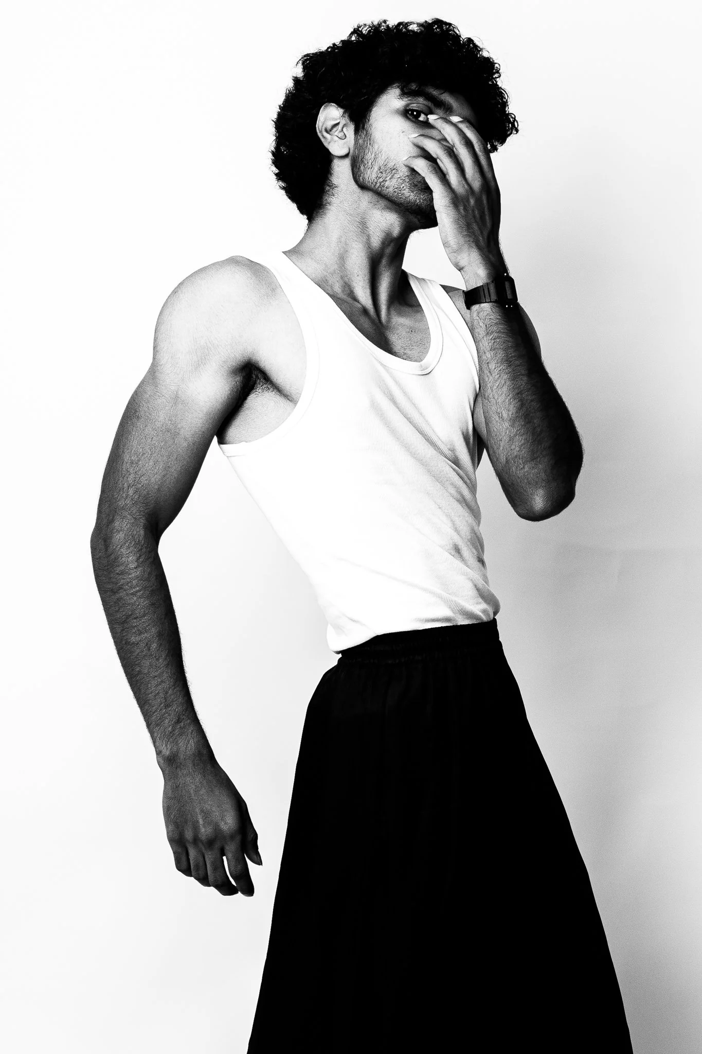 Black and white portrait of a man with curly hair wearing a white tank top, black pants, and a watch, with one hand covering part of his face.
