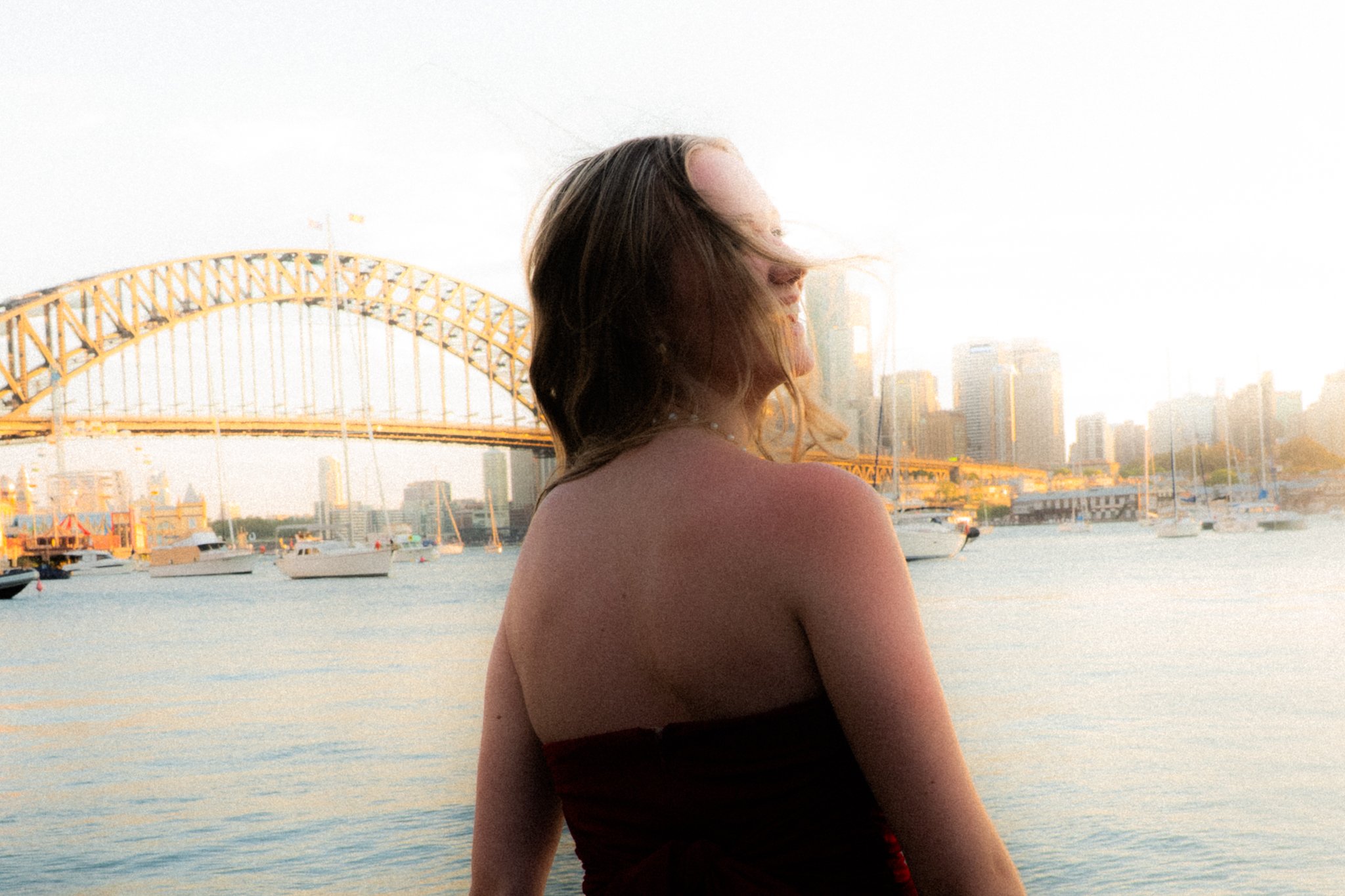 A woman with brown hair standing near a body of water with boats, a bridge, and a city skyline in the background during sunset.