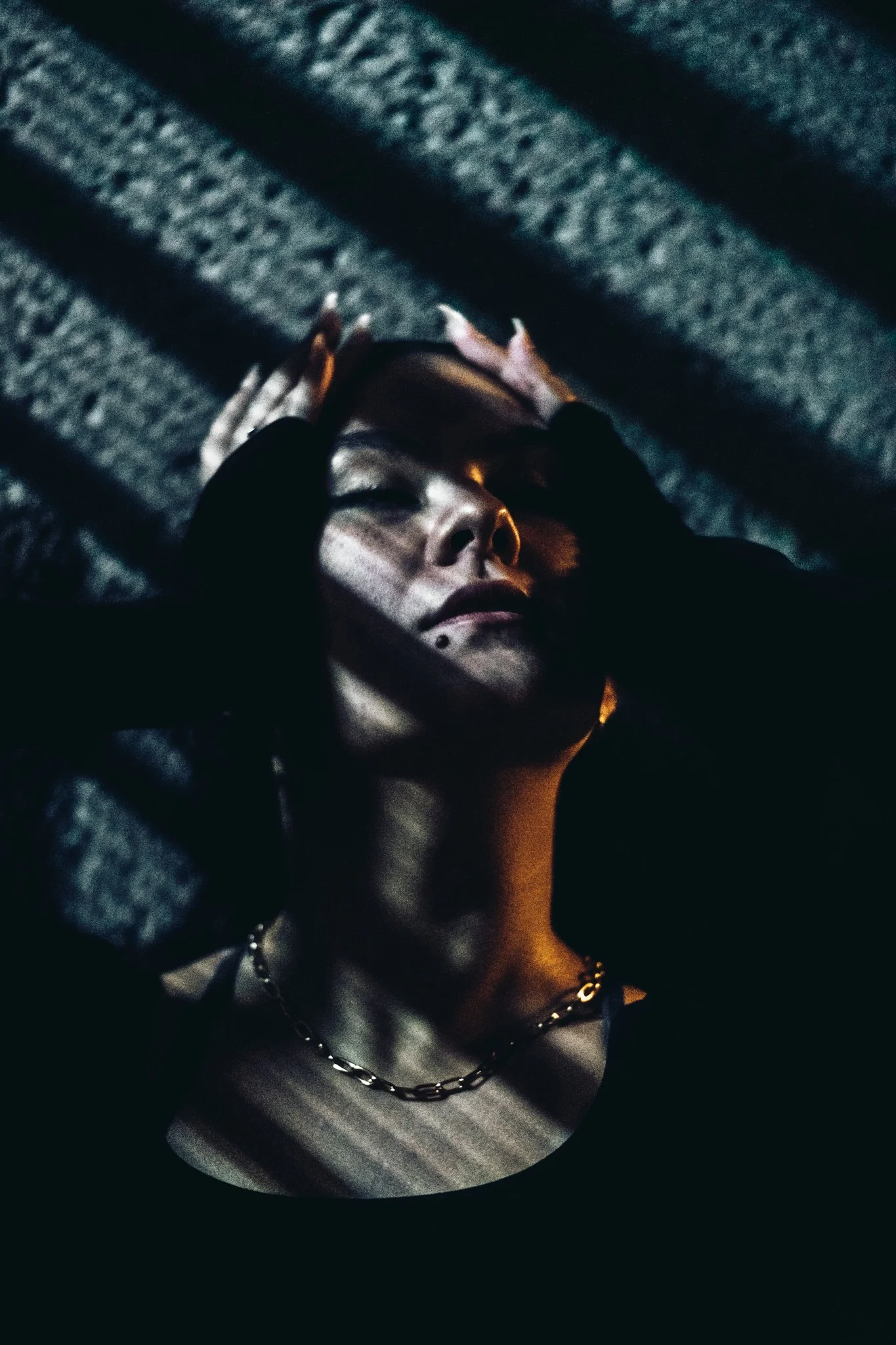 A woman with short hair and a small facial piercing, wearing a chain necklace and a black top, is lying down with her hands on her head. Shadows of blinds cast lines across her face. The background appears to be a textured surface, possibly a fabric 