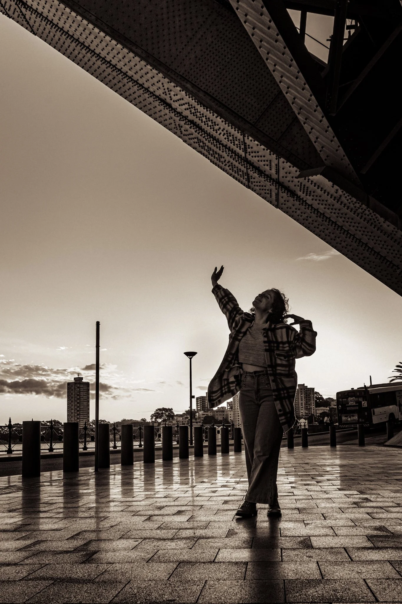 A woman standing outdoors on a paved walkway during sunset, striking a joyful pose with arms raised and one hand touching her head, with city buildings and a bus in the background.