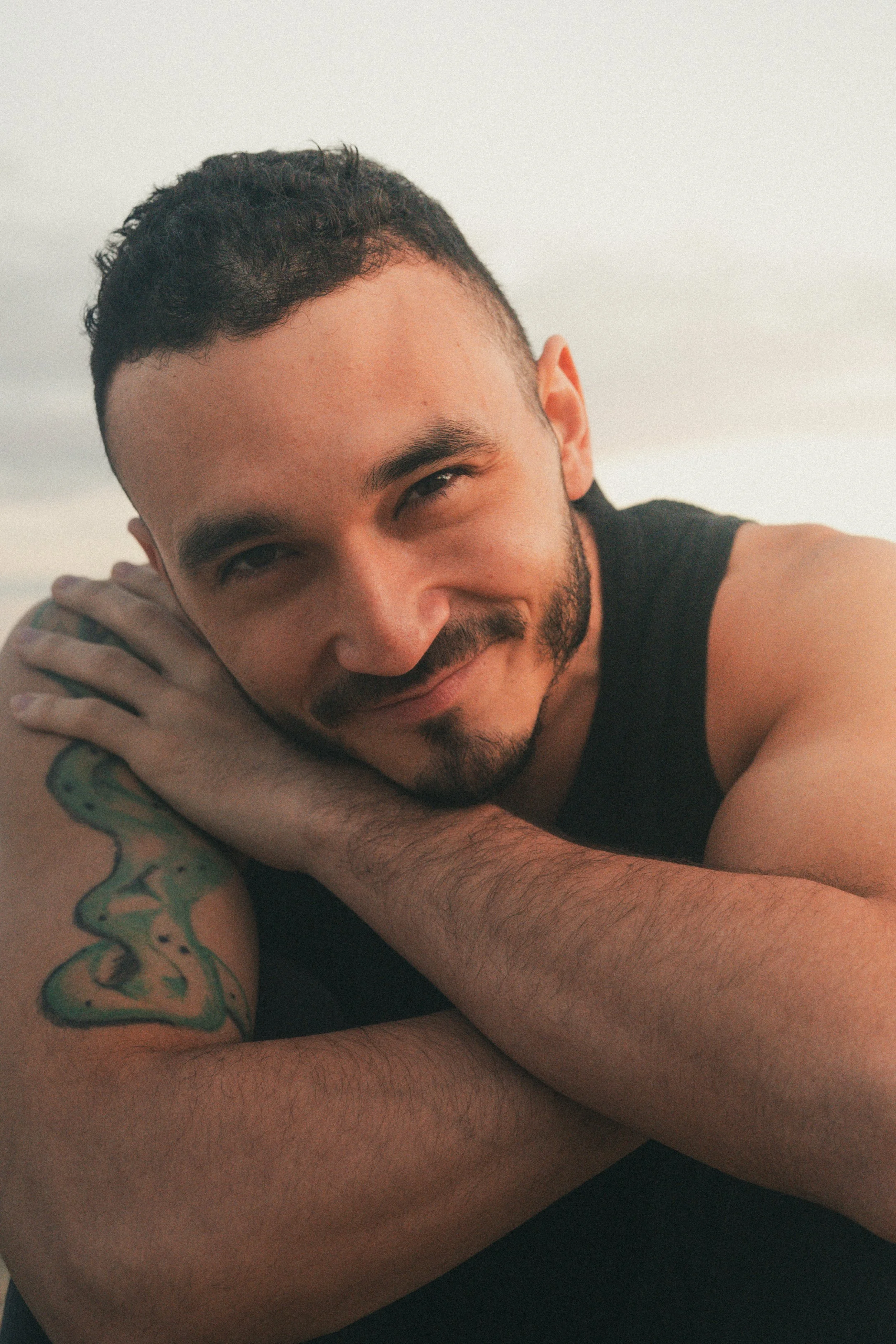 A young man with short dark hair and facial hair, smiling softly while resting his head on his folded arms. He has a tattoo on his right arm and is wearing a sleeveless black shirt.