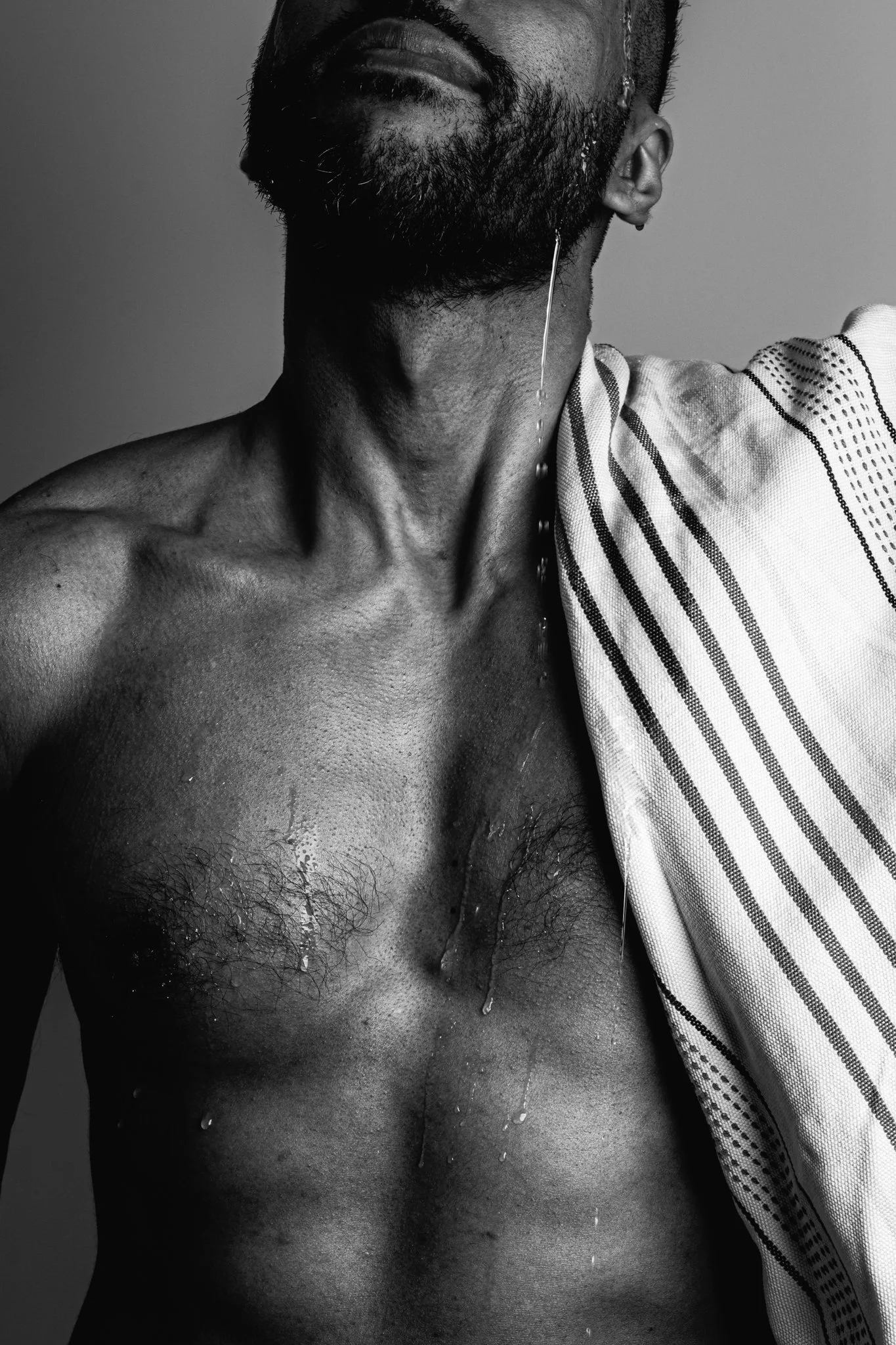Close-up black and white photograph of a man's partially visible face and upper body, with droplets of water on his skin, holding a striped cloth.