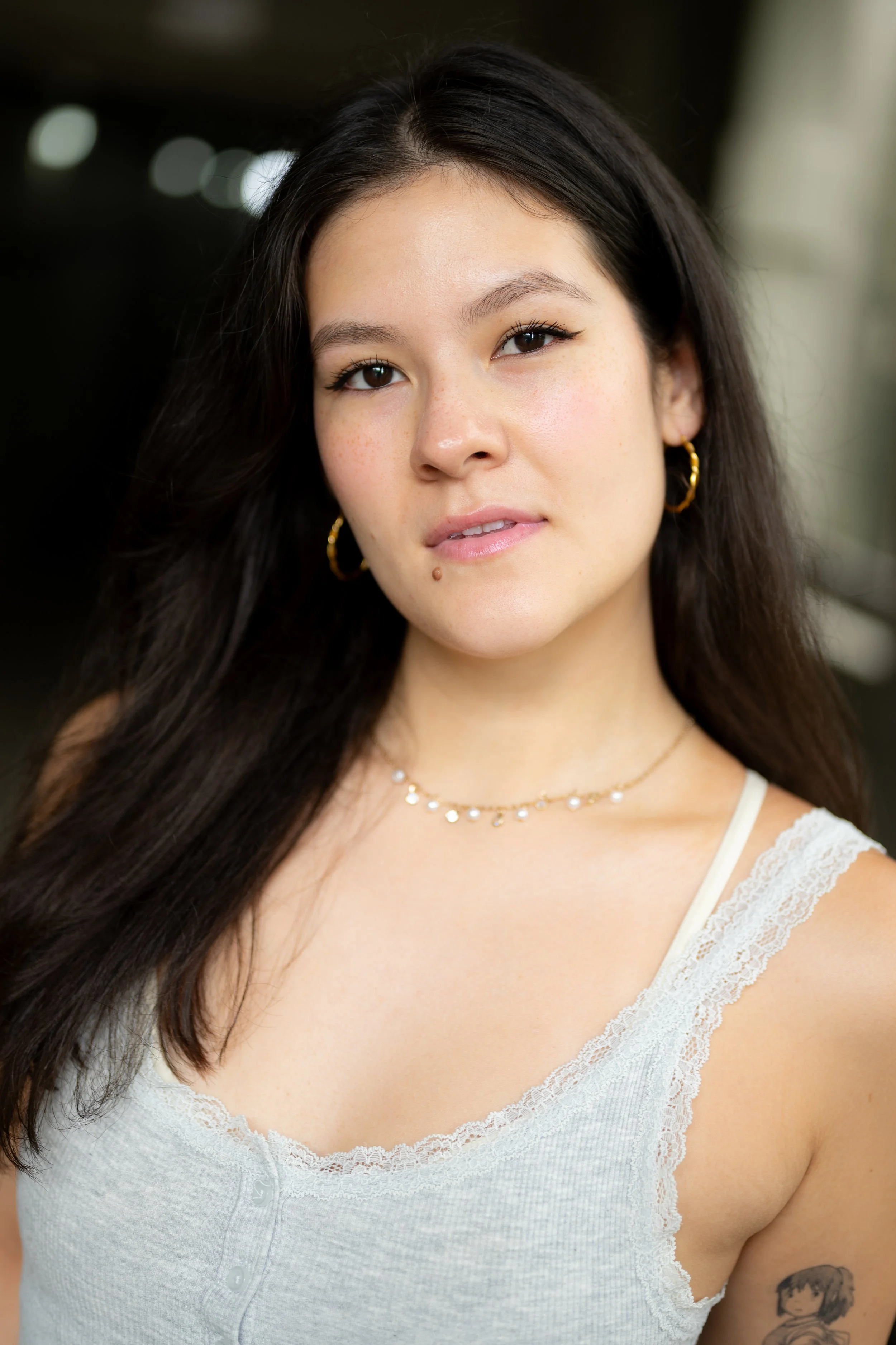 Close-up portrait of a young woman with long dark hair, wearing a white lace-trimmed top, gold hoop earrings, a delicate necklace, and a small tattoo on her arm.
