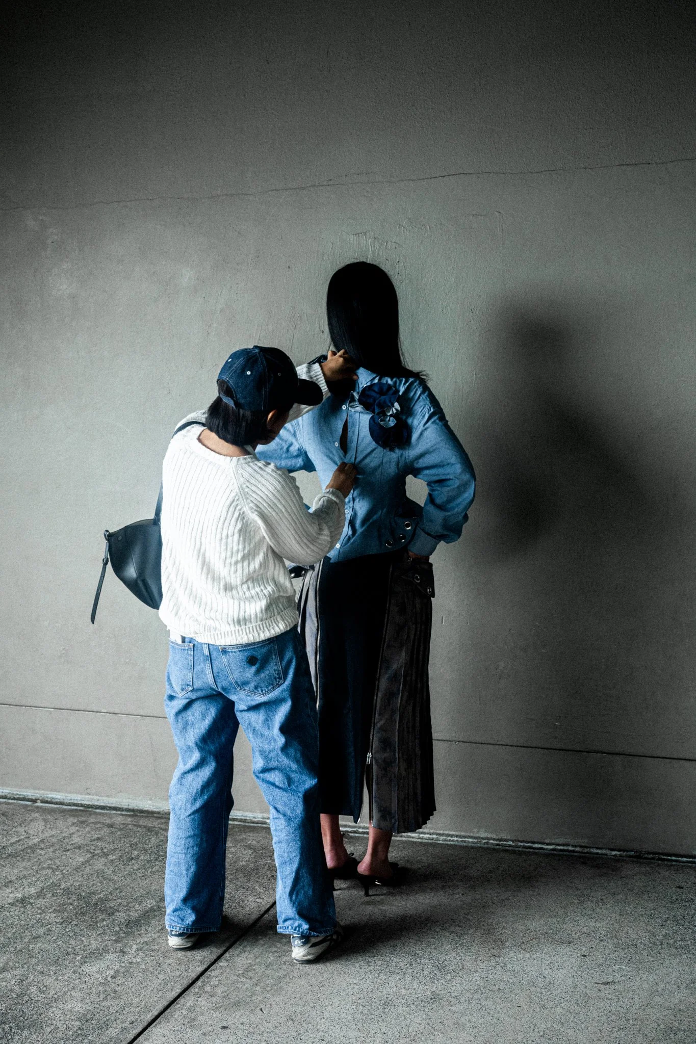 Two women standing against a plain wall, one wearing a white sweater and blue jeans, the other in a blue jacket and black skirt, with the woman in the blue jacket adjusting her hair.