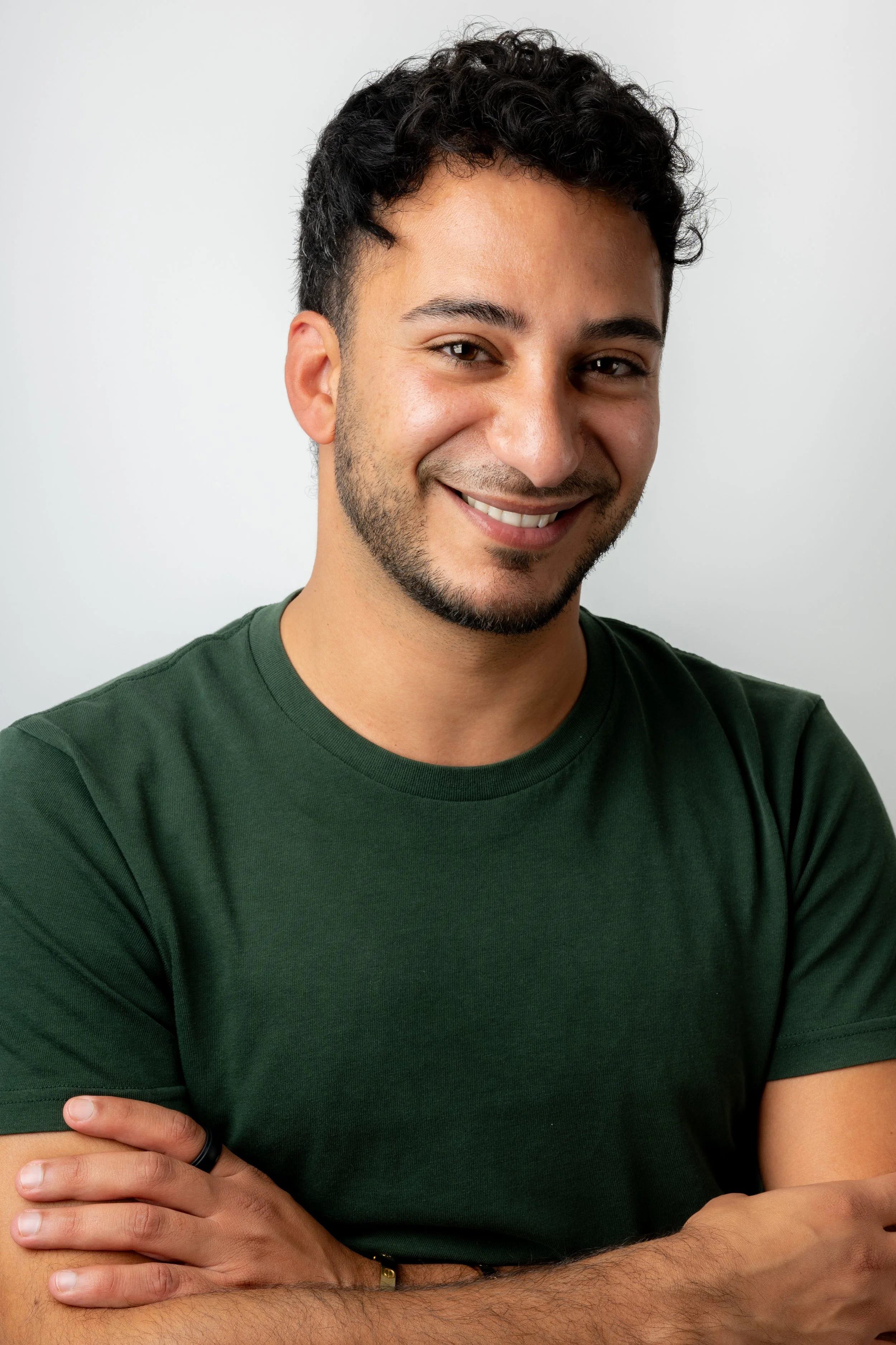 A young man with curly dark hair, a beard, and a friendly smile, wearing a dark green T-shirt, standing against a plain white background.