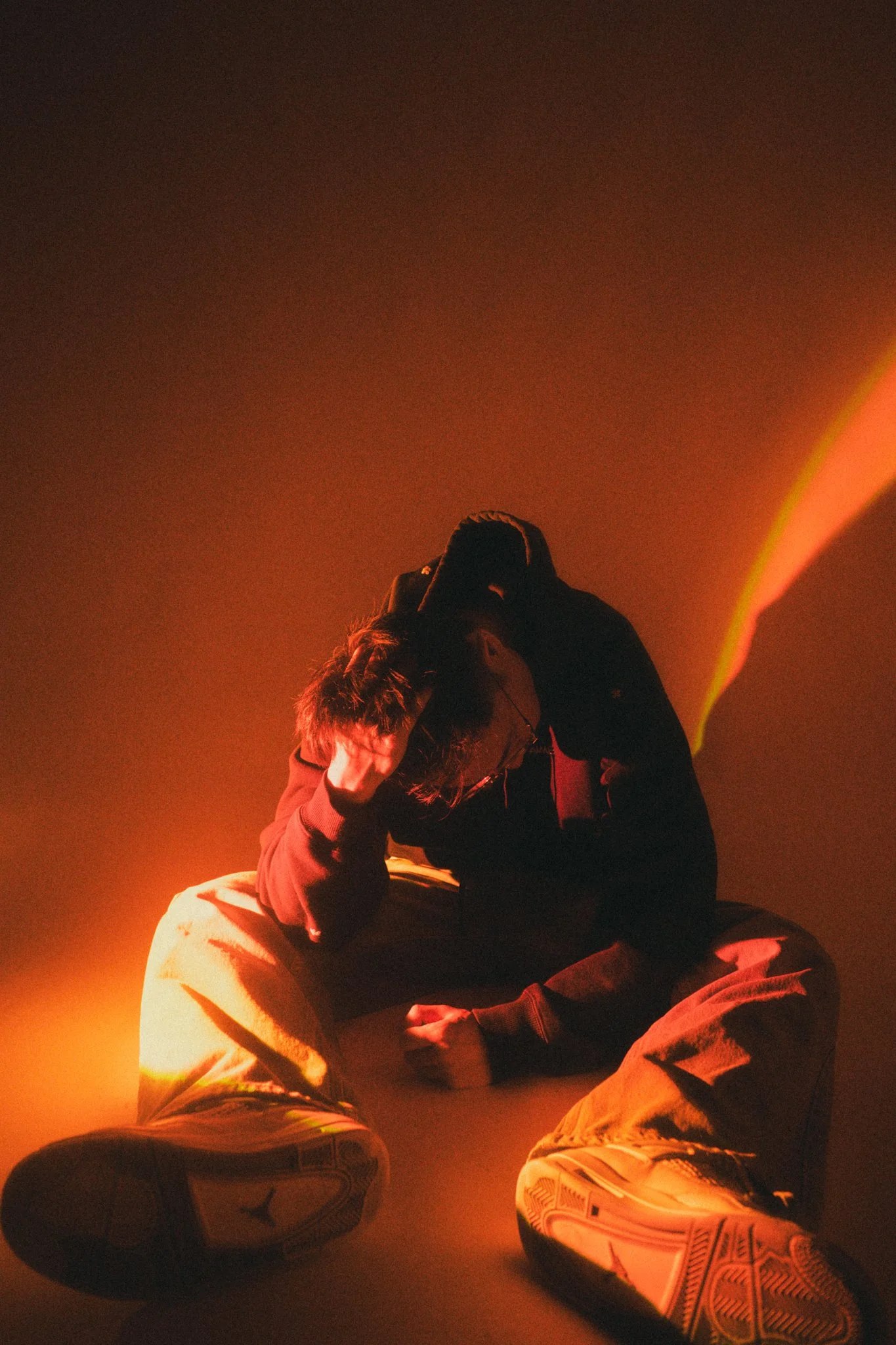 A person sits on the floor with their head down and hand on their forehead, illuminated by warm orange and yellow lighting.