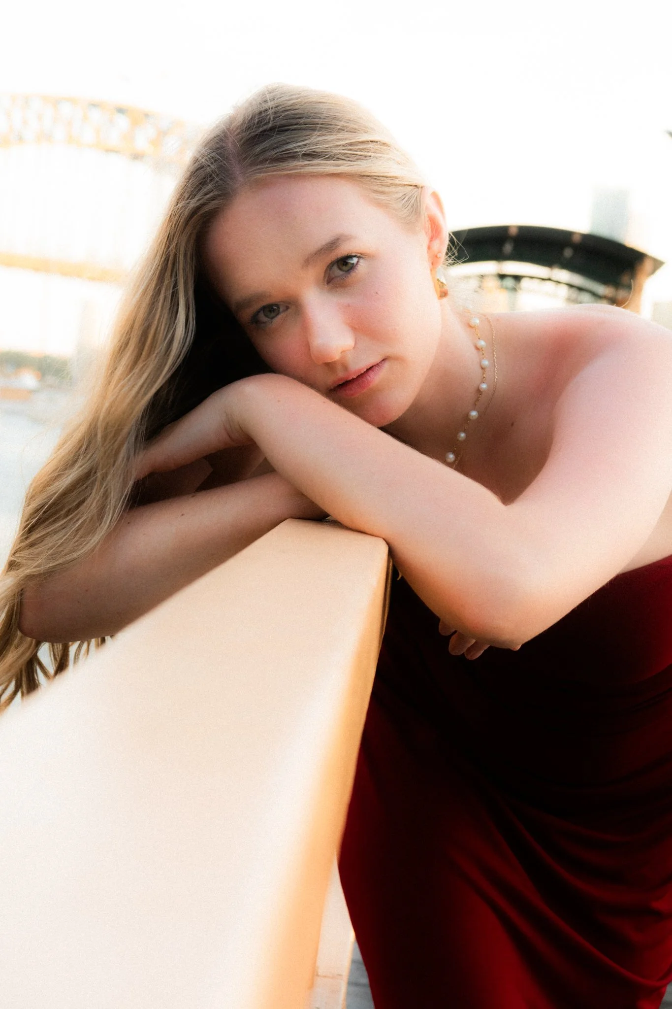 A young woman with long blonde hair leaning on a beige railing outdoors, wearing a maroon dress and jewelry, looking at the camera with a neutral expression.