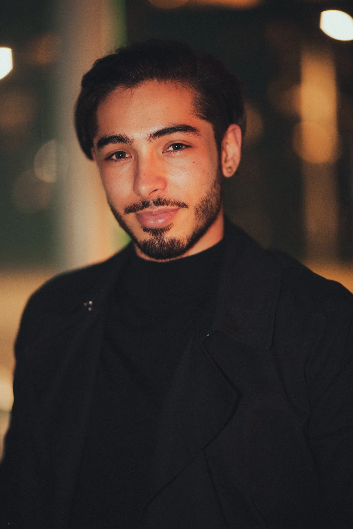 A young man with dark hair, beard, and earrings, wearing a black jacket and turtleneck, smiling at the camera in a dimly lit indoor setting with blurred background lights.