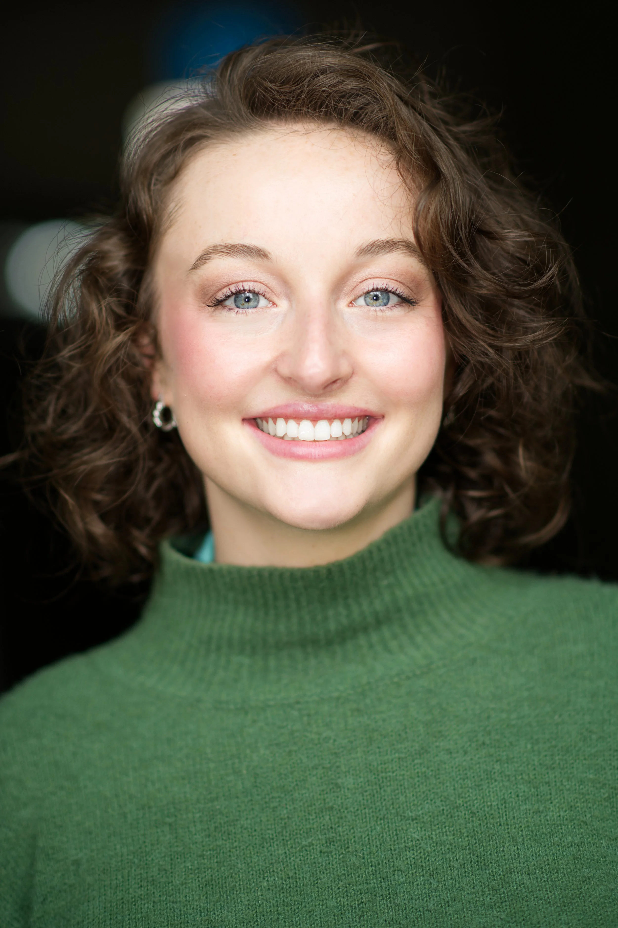 A smiling woman with curly brown hair, blue eyes, and wearing a green sweater.