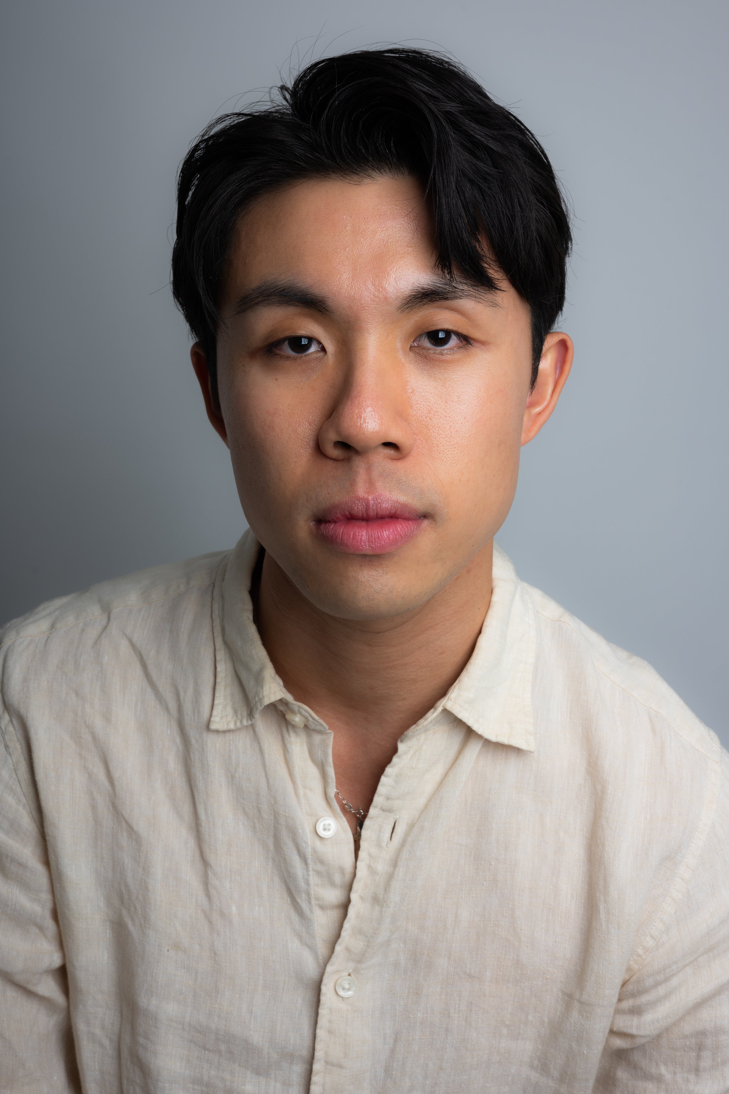 Close-up portrait of a young Asian man with black hair wearing a light-colored button-up shirt against a gray background.