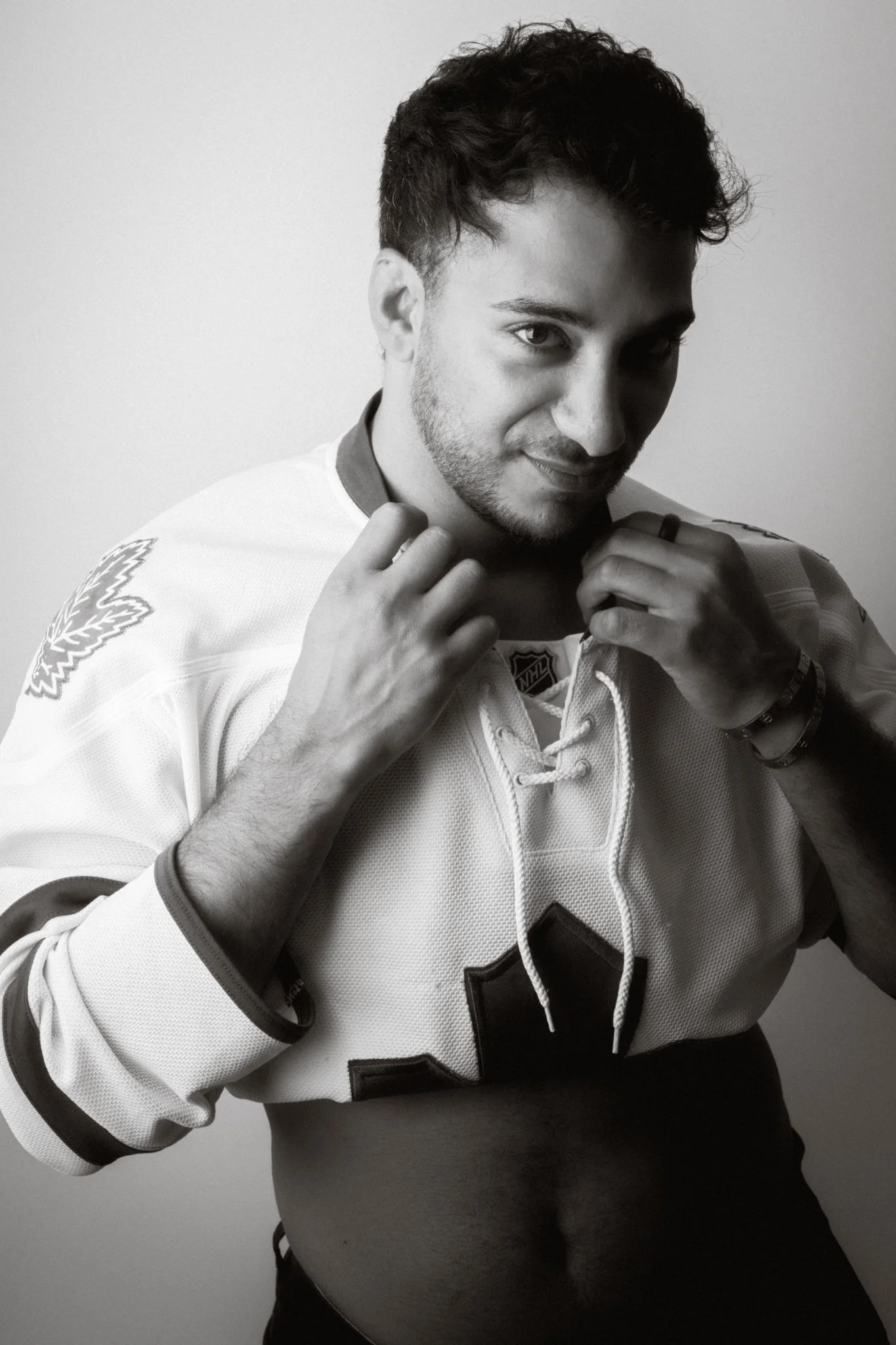 Black and white photo of a young man lifting the collar of his hockey jersey, revealing his abdomen. He has short, dark, curly hair, a beard, and is looking directly at the camera with a slight smile.