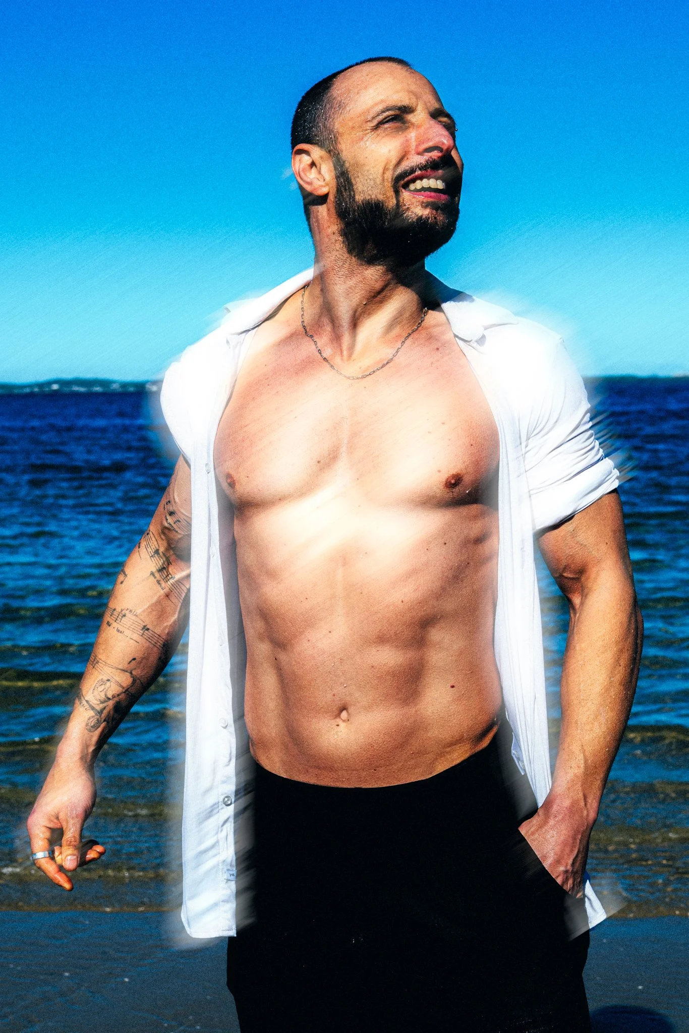Man with a beard and tattoos standing shirtless at the beach, with a blue sky and ocean in the background.