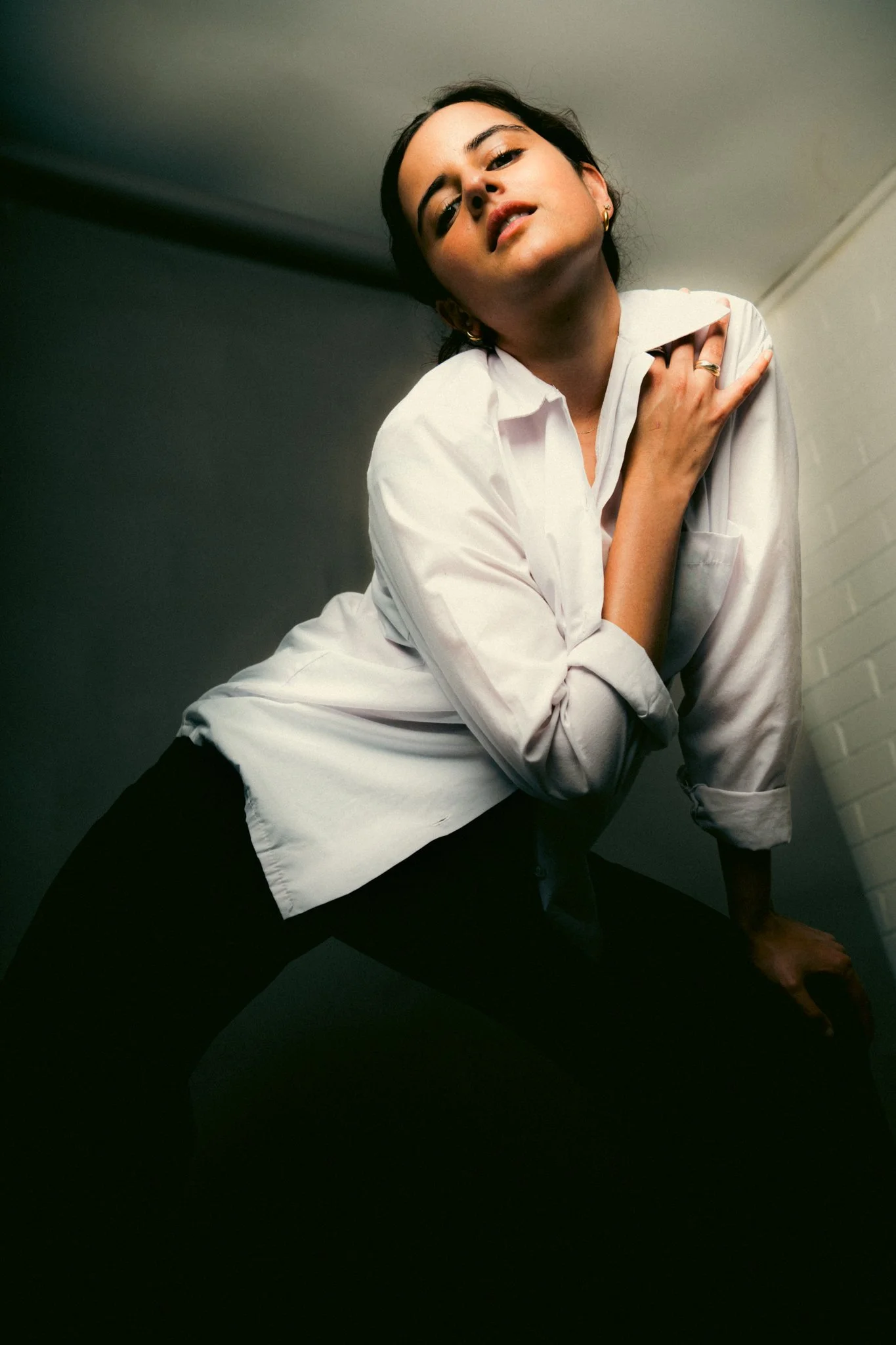 A woman wearing a white shirt and black pants posing in a dimly lit room with a plain background.
