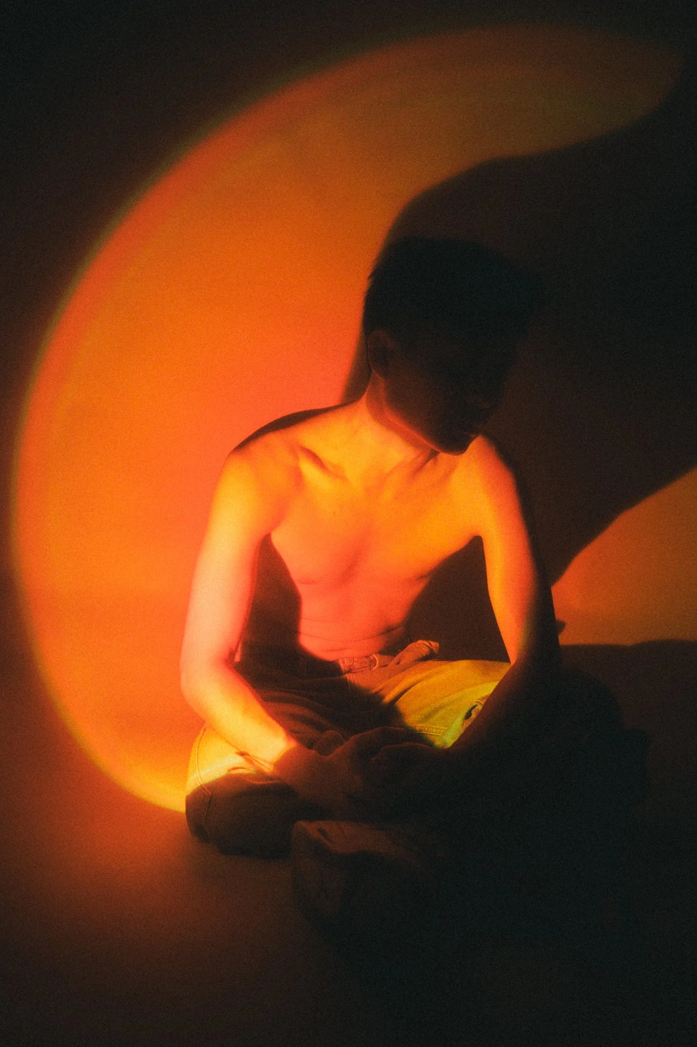 A shirtless man sitting cross-legged in a dimly lit room with warm orange lighting, with his head bowed and shadowed face.