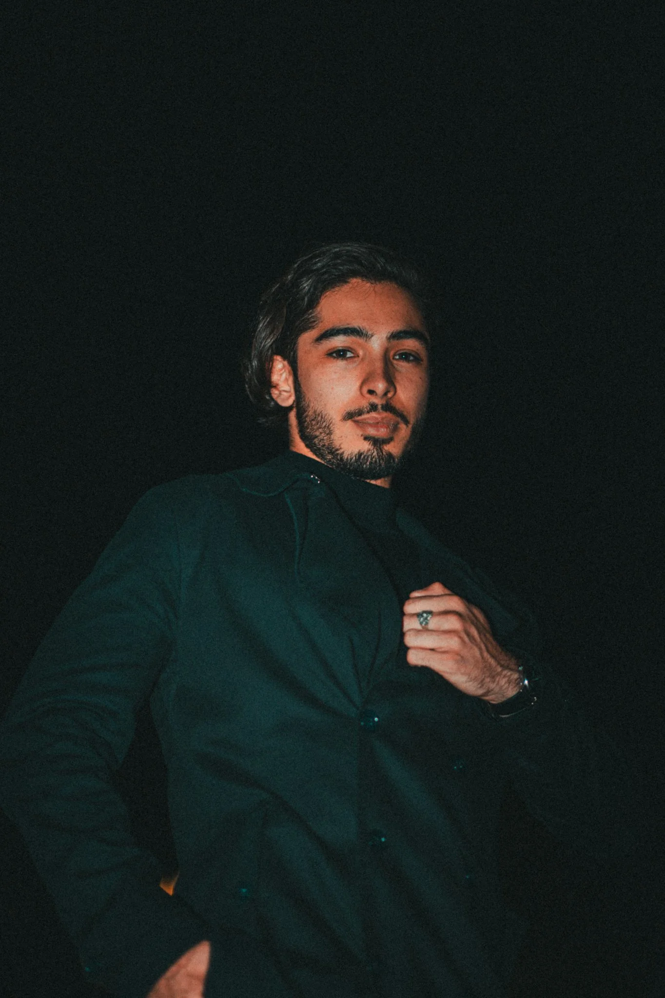 A man with dark hair and a beard, wearing a black shirt and jacket, looking into the camera against a dark background.
