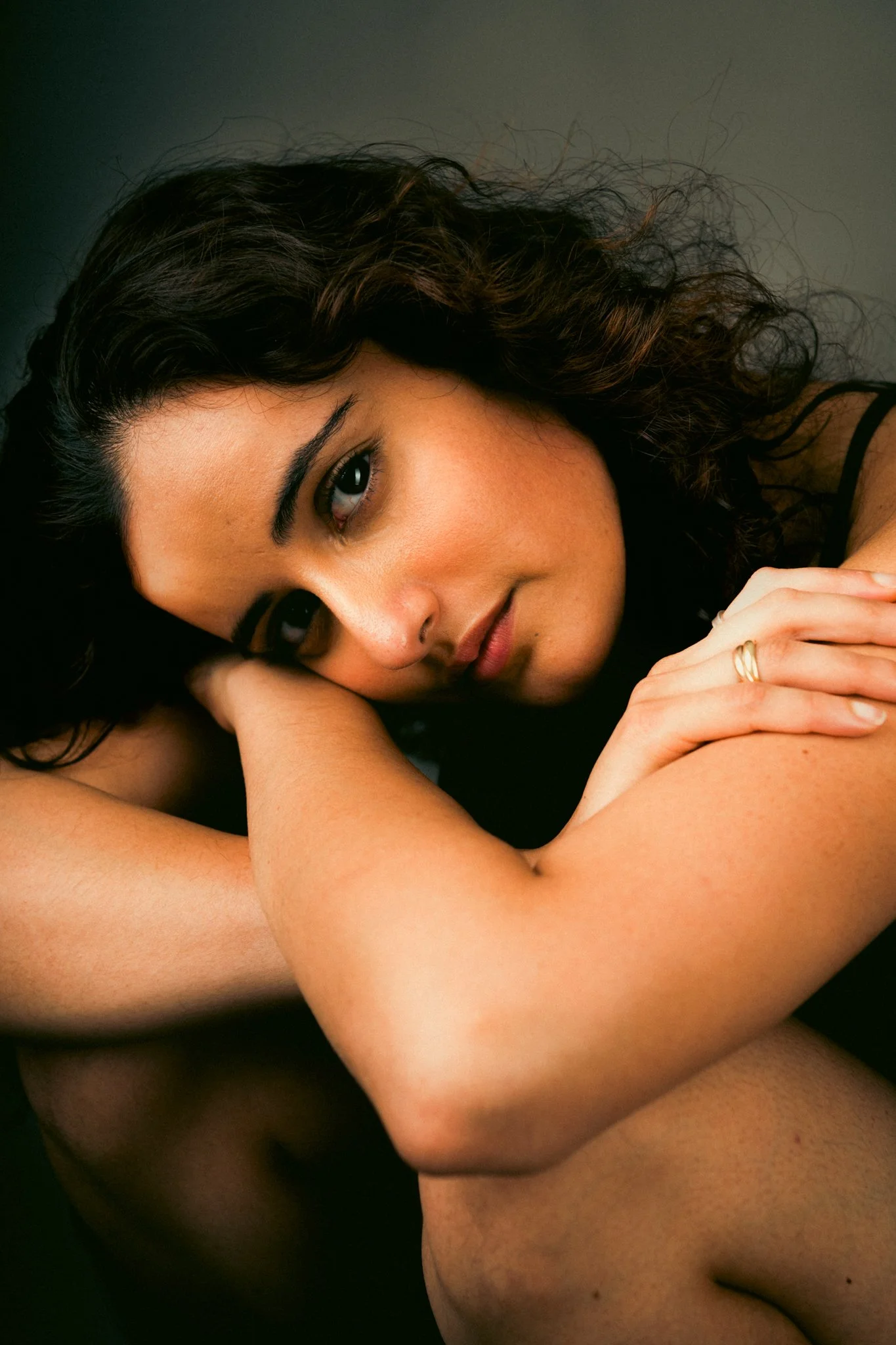 A woman with curly dark hair resting her head on her arm, looking at the camera with a gentle expression.