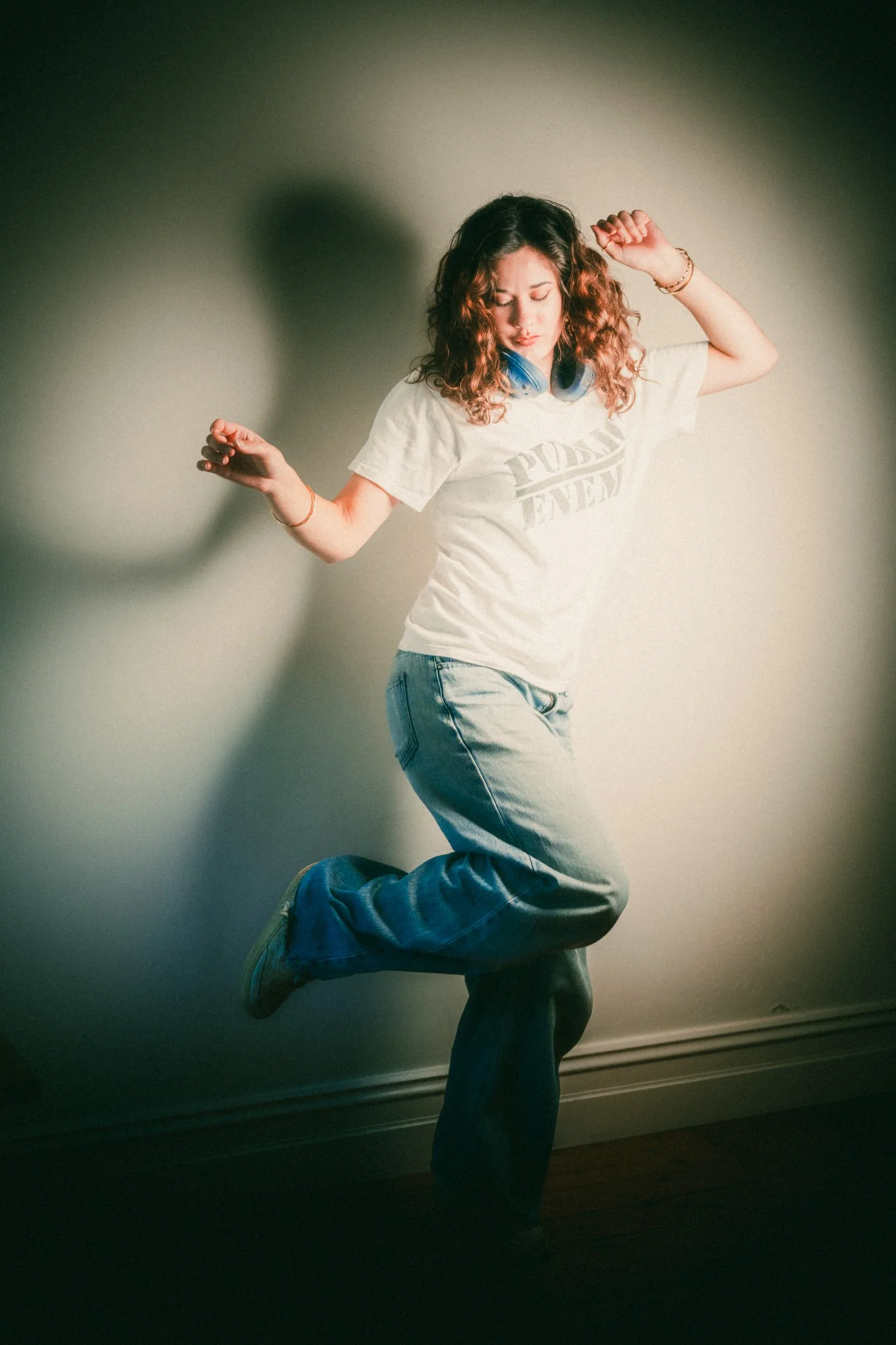 A young woman with curly hair is dancing with her eyes closed, wearing a white T-shirt and blue jeans, with a beige wall in the background.