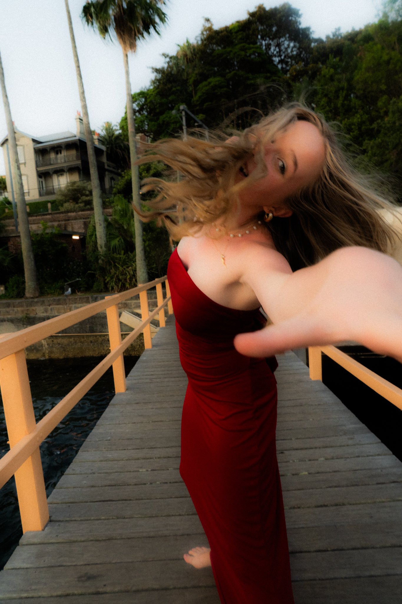 A woman in a red dress standing on a wooden dock by the water, reaching out towards the camera, with her hair blowing in the wind, during sunset.