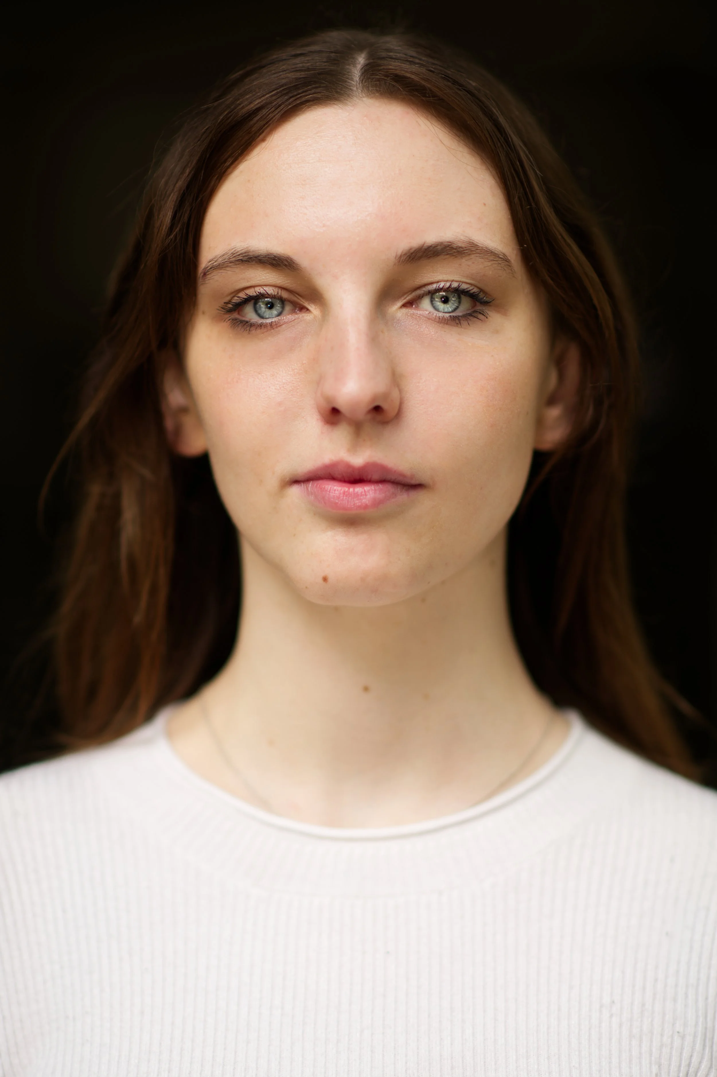 A young woman with blue eyes, light skin, and brown hair wearing a white top against a dark background.