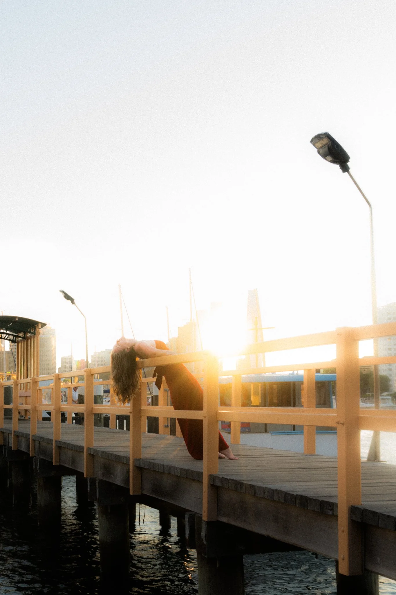 Woman with long hair in a red dress leaning backwards on a yellow railing of a wooden pier at sunset, overlooking water and city skyline.