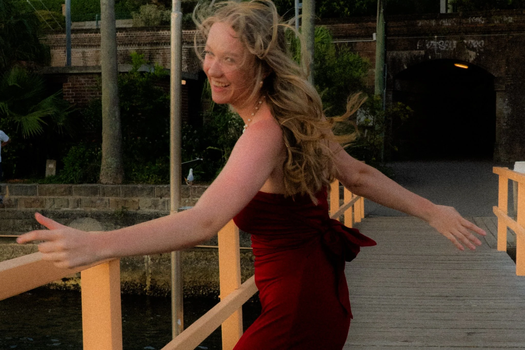 Woman with long blonde hair in a red dress standing on a wooden dock near water, smiling and twirling with arms outstretched.