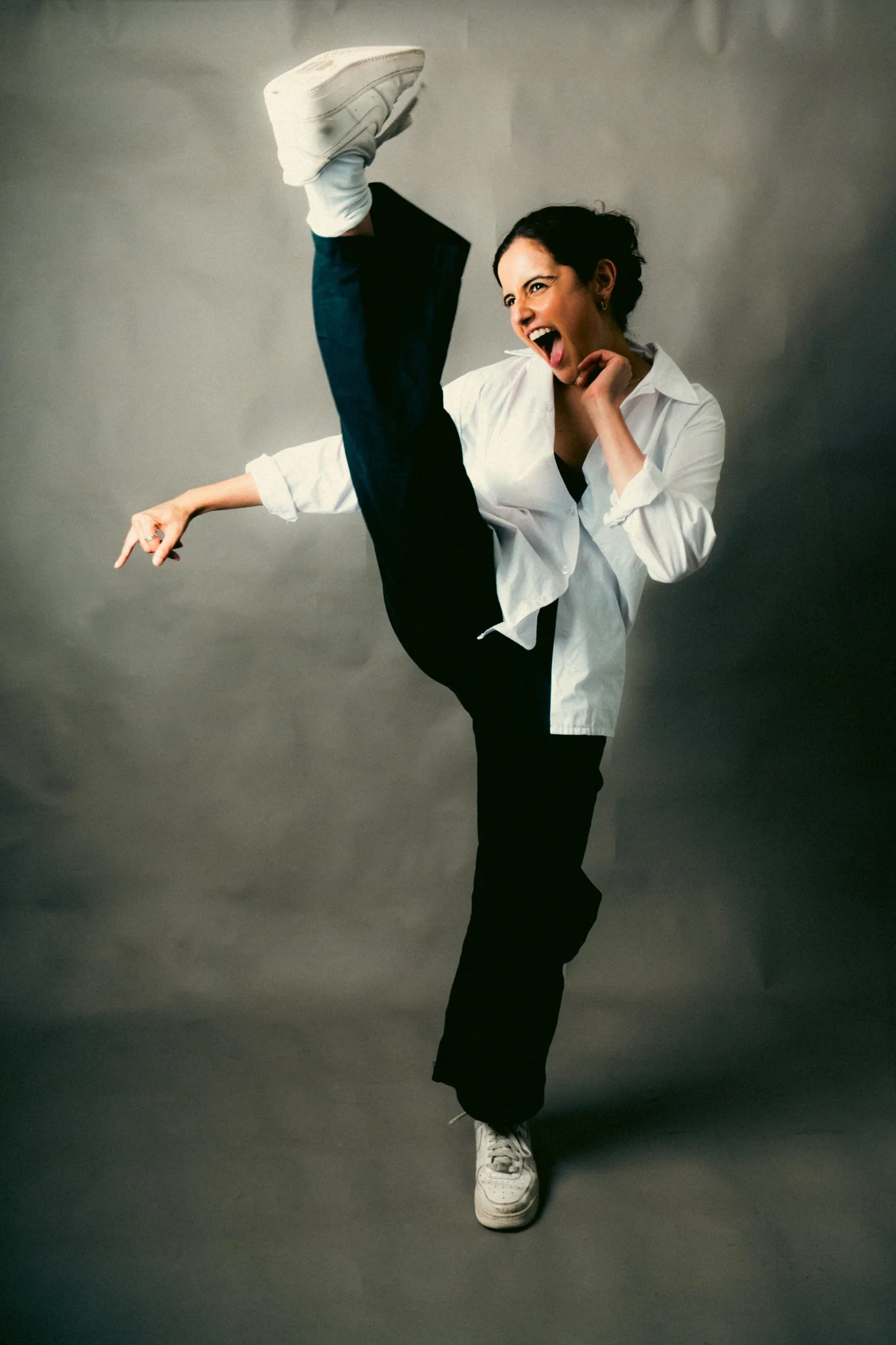 Woman in white shirt and black pants performing a high kick, smiling and pointing.