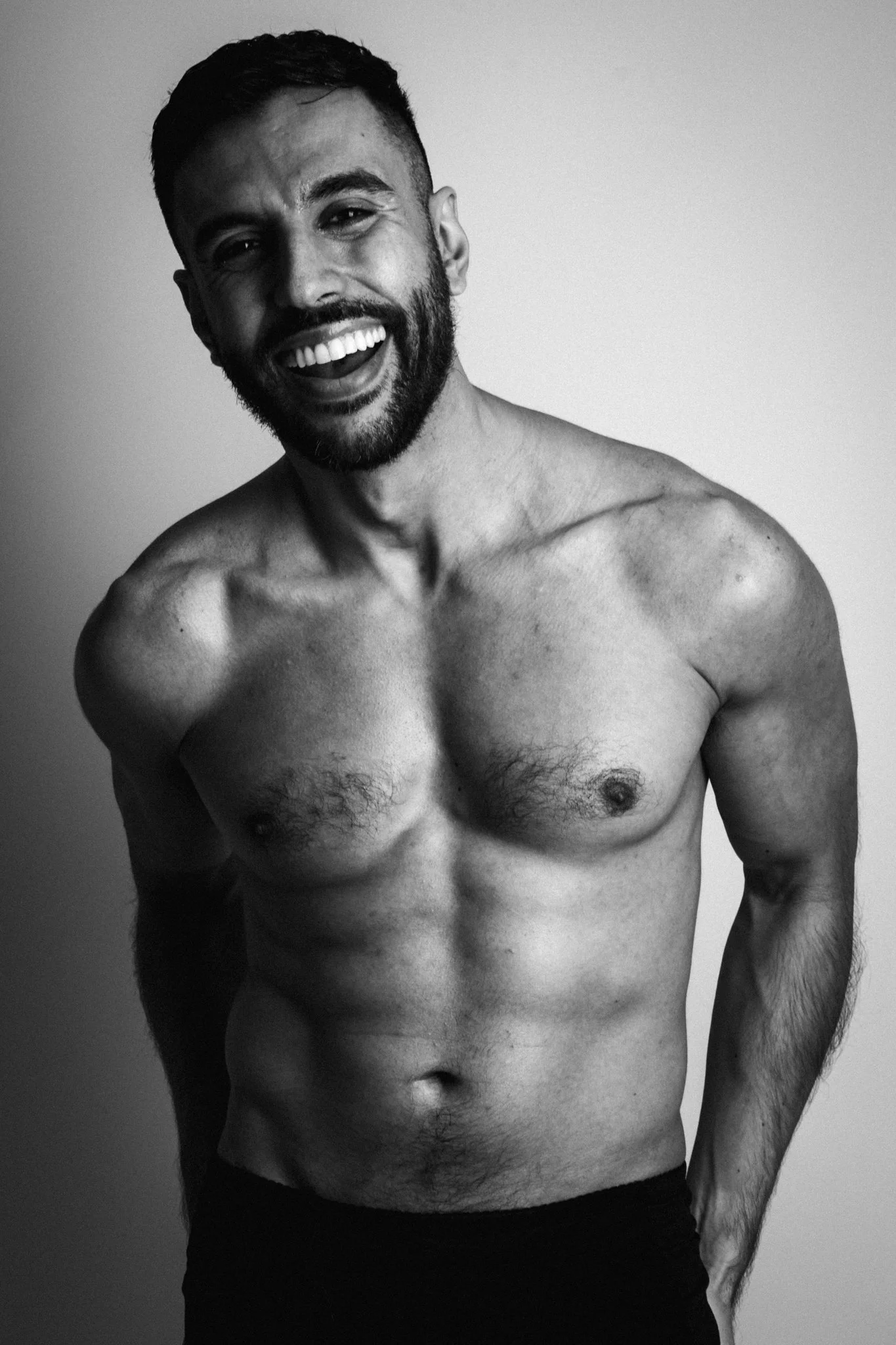 Black-and-white photo of a shirtless man with a beard, smiling and looking at the camera, his head slightly tilted, with a plain background.