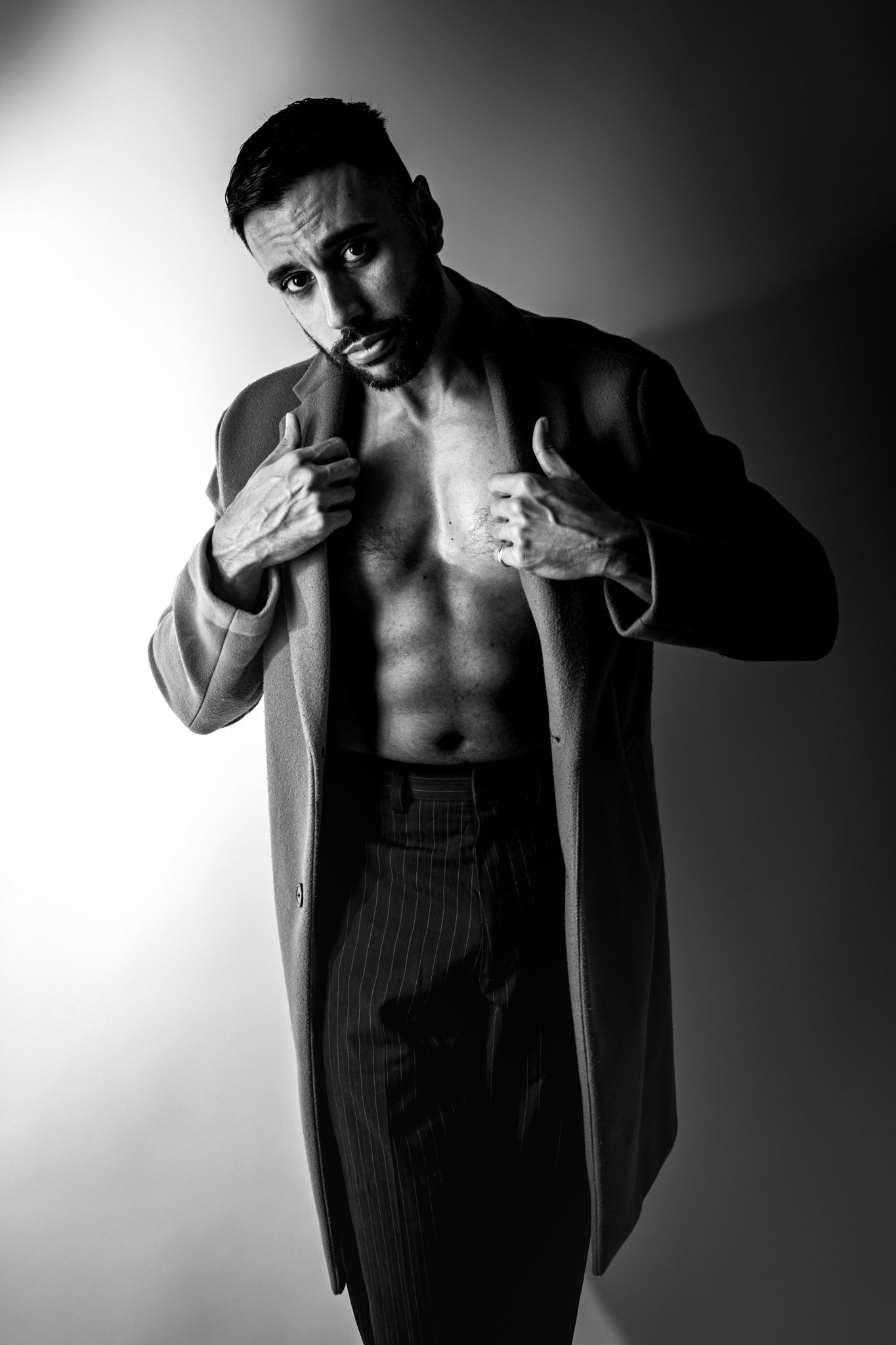 Black and white photo of a shirtless man with a beard, wearing a blazer and pinstriped pants, adjusting his blazer on a plain background.