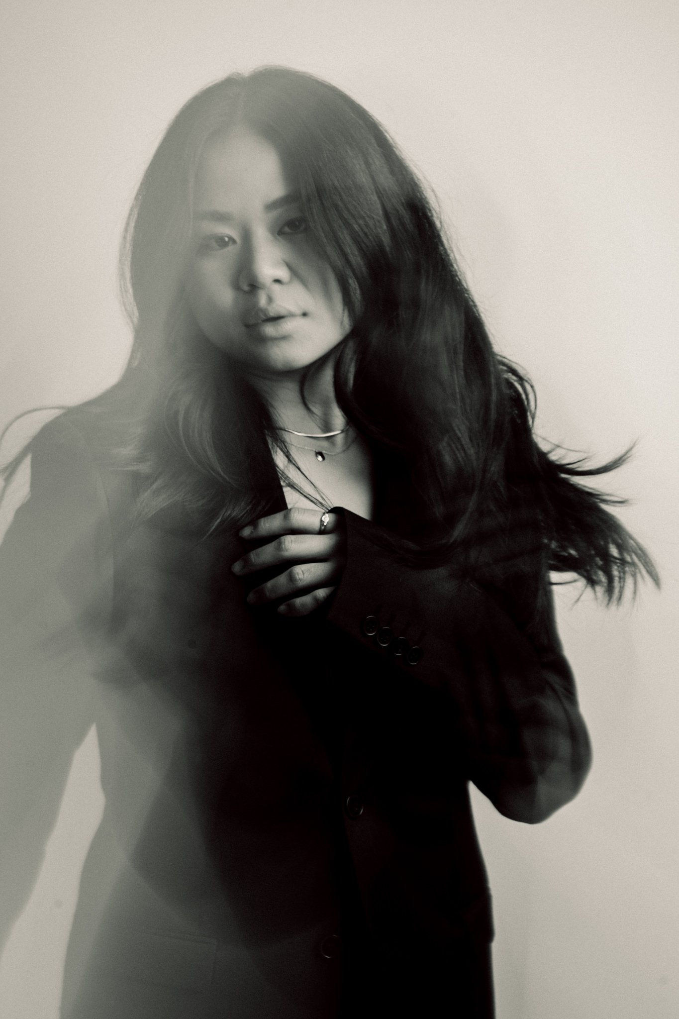 A black and white portrait of a young woman with long, dark hair, wearing a blazer, layered necklaces, and rings, posing against a plain background.