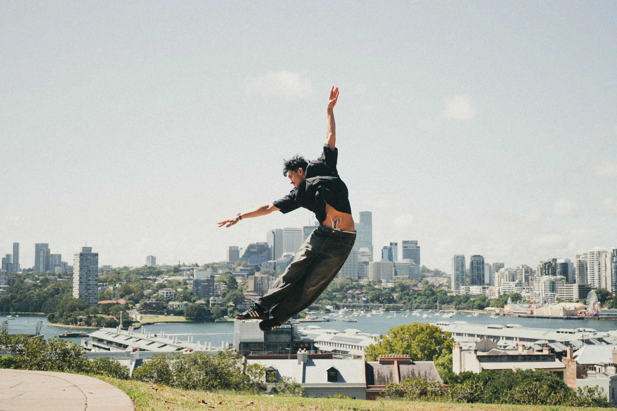 A person mid-air in a park with a city skyline in the background, with water and buildings visible beyond trees and rooftops.