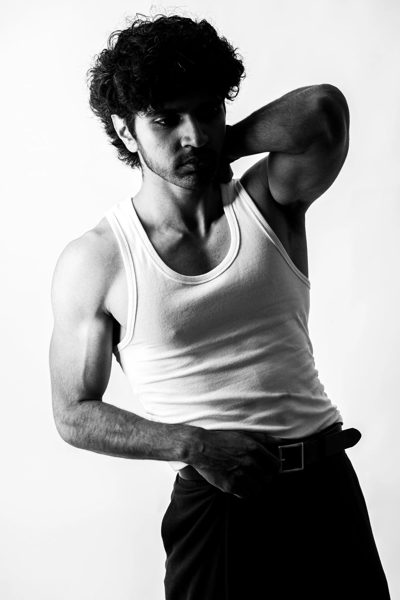 A black and white photo of a man with curly hair, wearing a white tank top, with one arm behind his head and looking to the side.