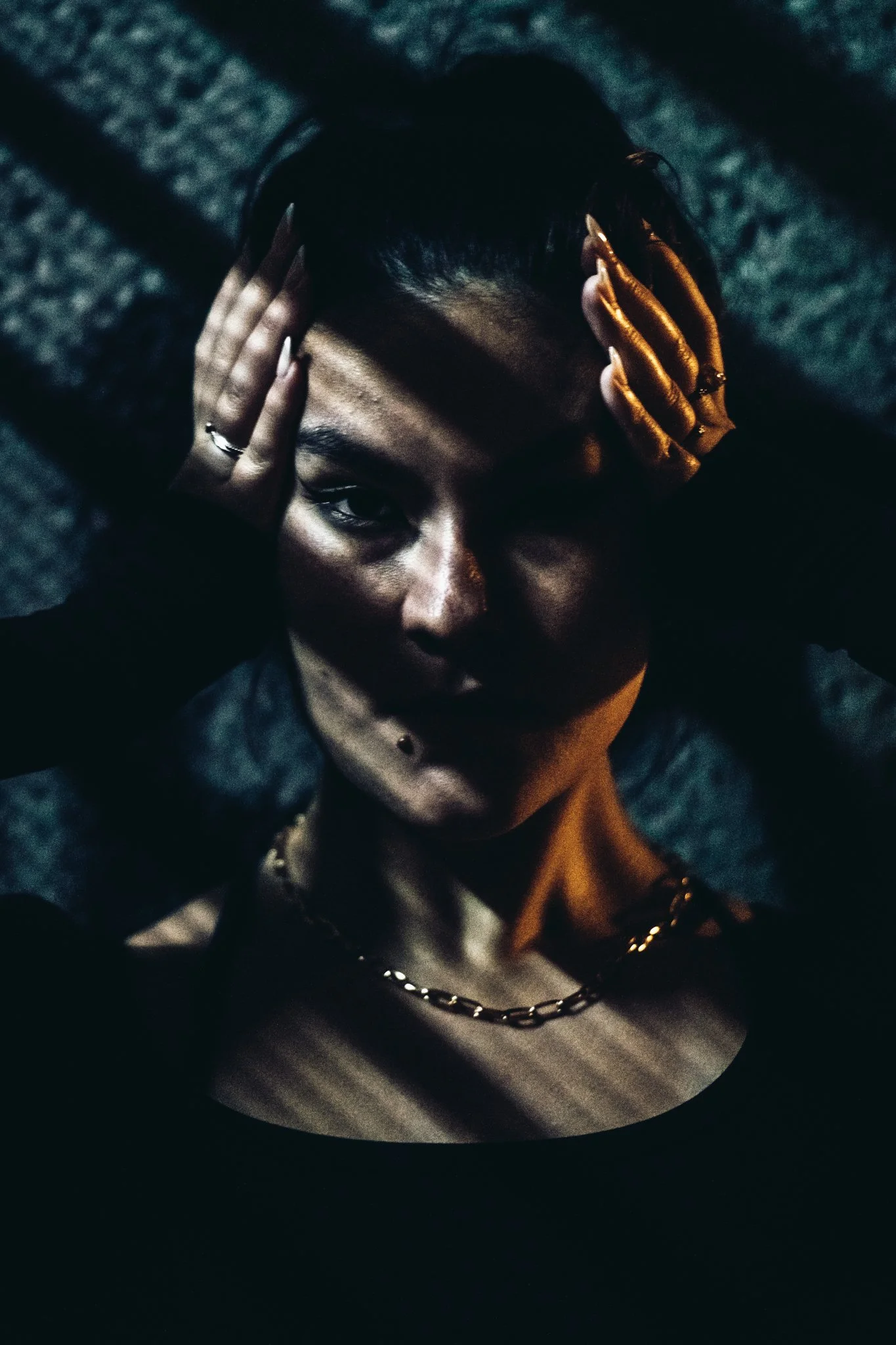 A dark, moody portrait of a woman with short dark hair, wearing a black top and gold accessories, holding her hands to her head with shadowed lighting.