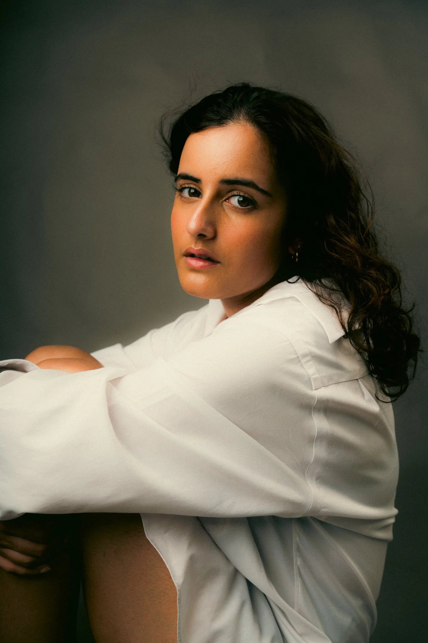Portrait of a woman with dark curly hair, wearing a white long-sleeve shirt, looking at the camera against a neutral background.
