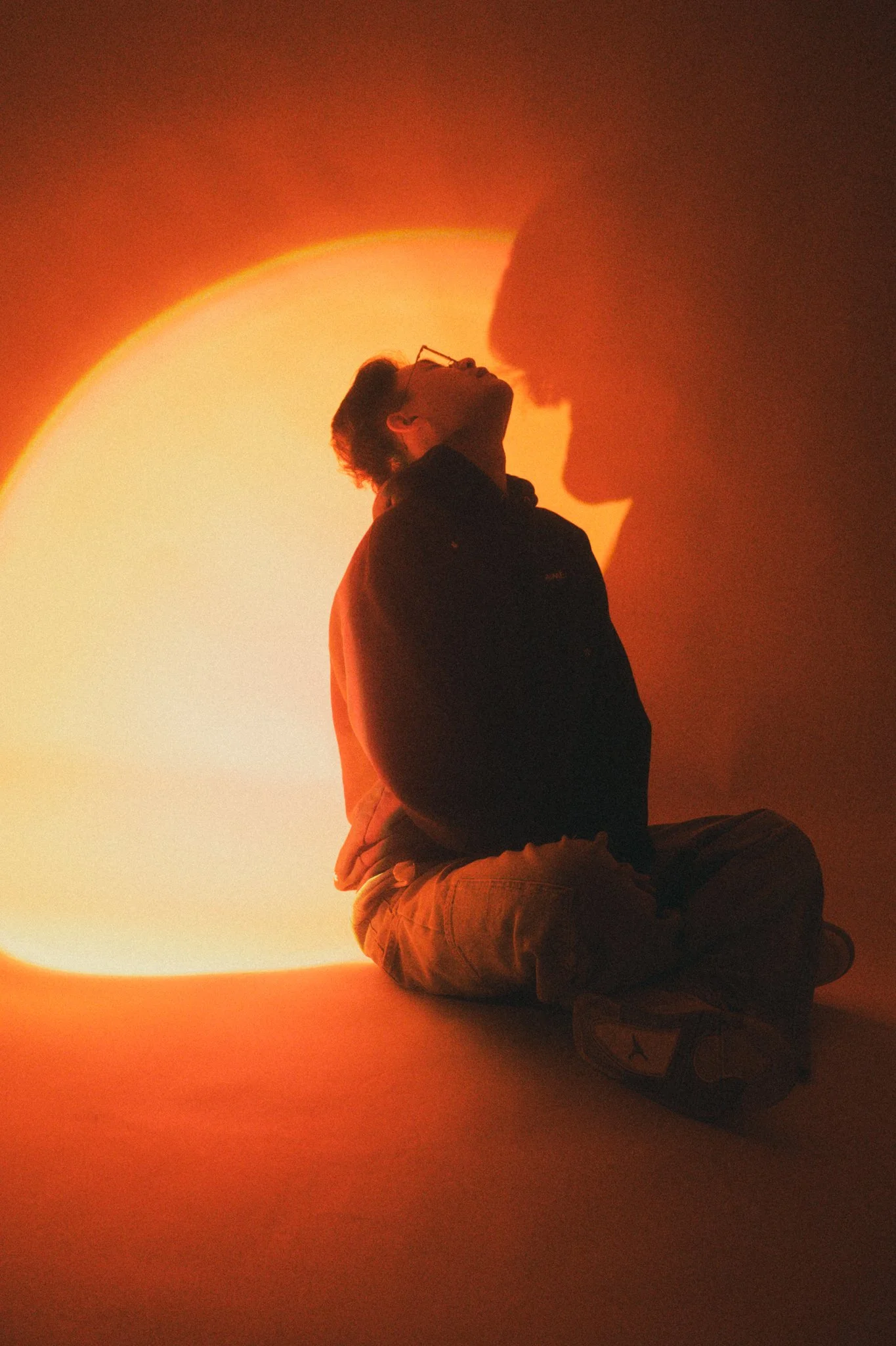 A person sitting cross-legged on the floor with their head tilted back, facing a warm, glowing light source that creates a large shadow of another person's face on the wall behind them.