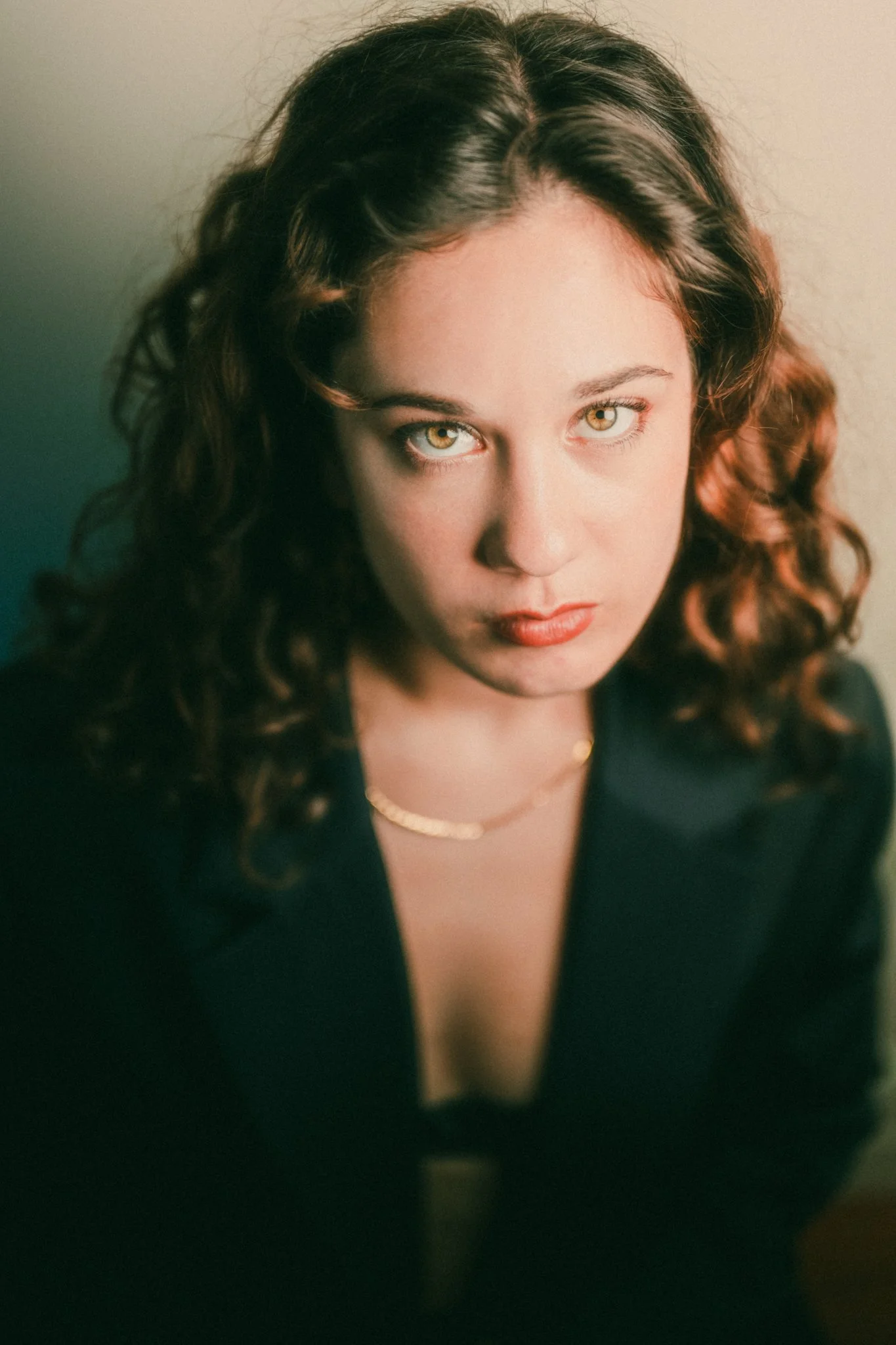 Close-up of a woman with curly brown hair, wearing a black blazer and gold necklace, looking directly at the camera.