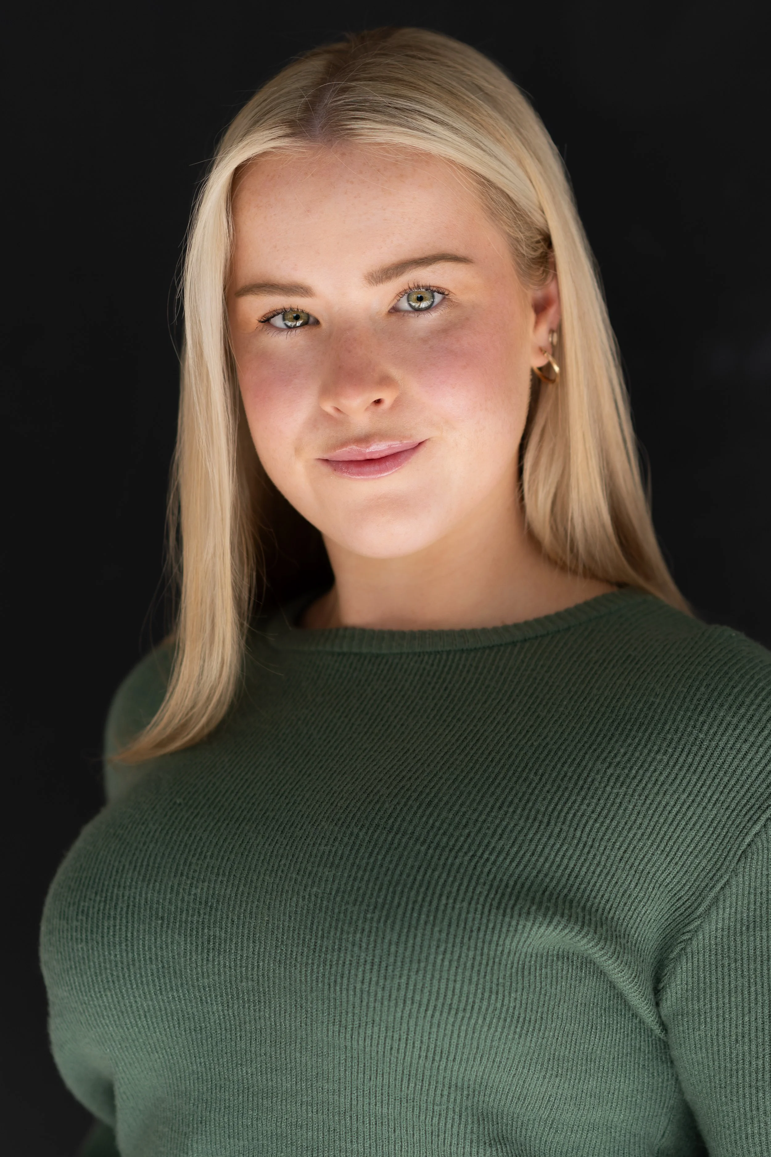 Close-up portrait of a young woman with long blonde hair, wearing a green sweater, looking at the camera with a slight smile, against a black background.