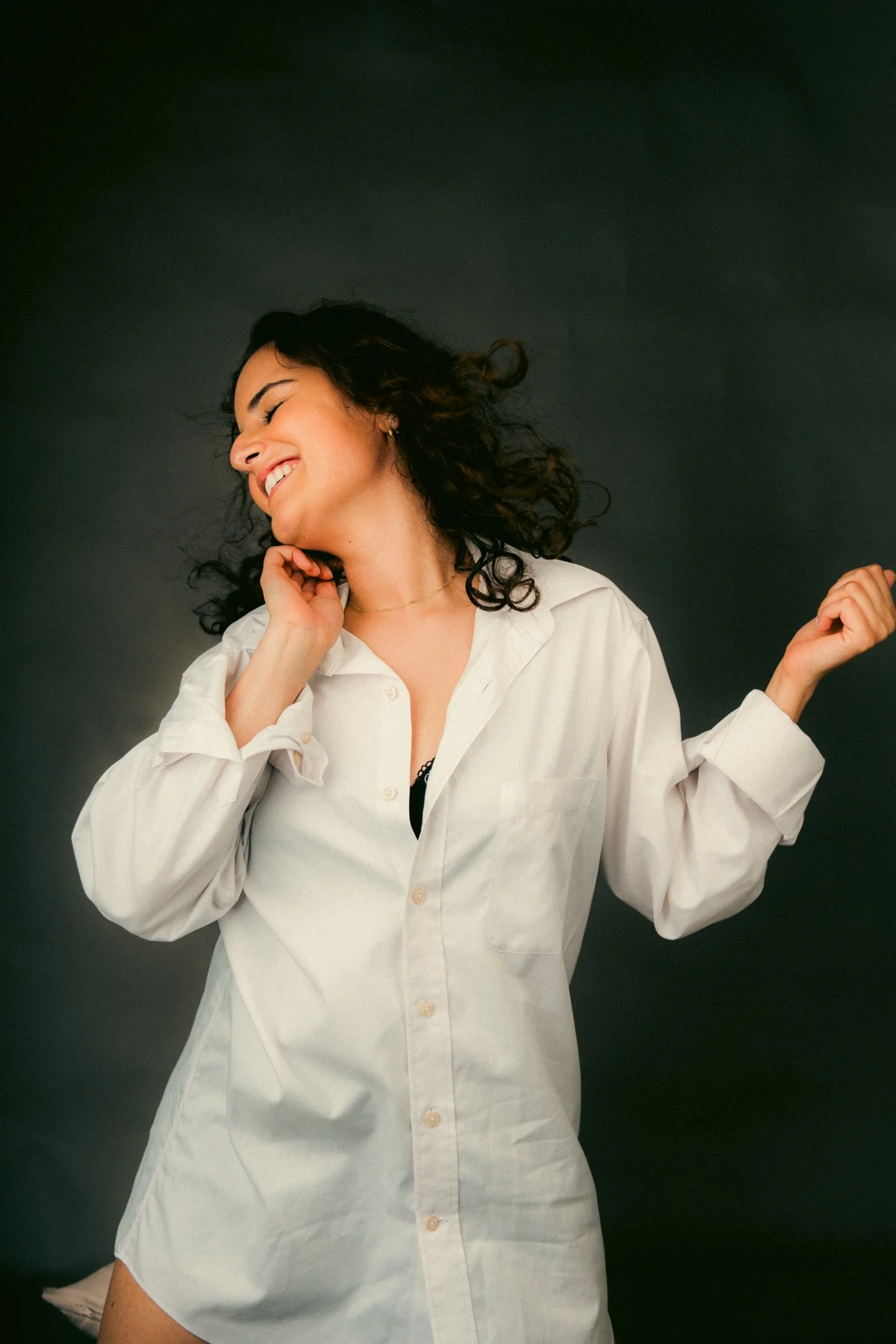 A woman with curly hair wearing a white button-down shirt, smiling with eyes closed, against a dark background.